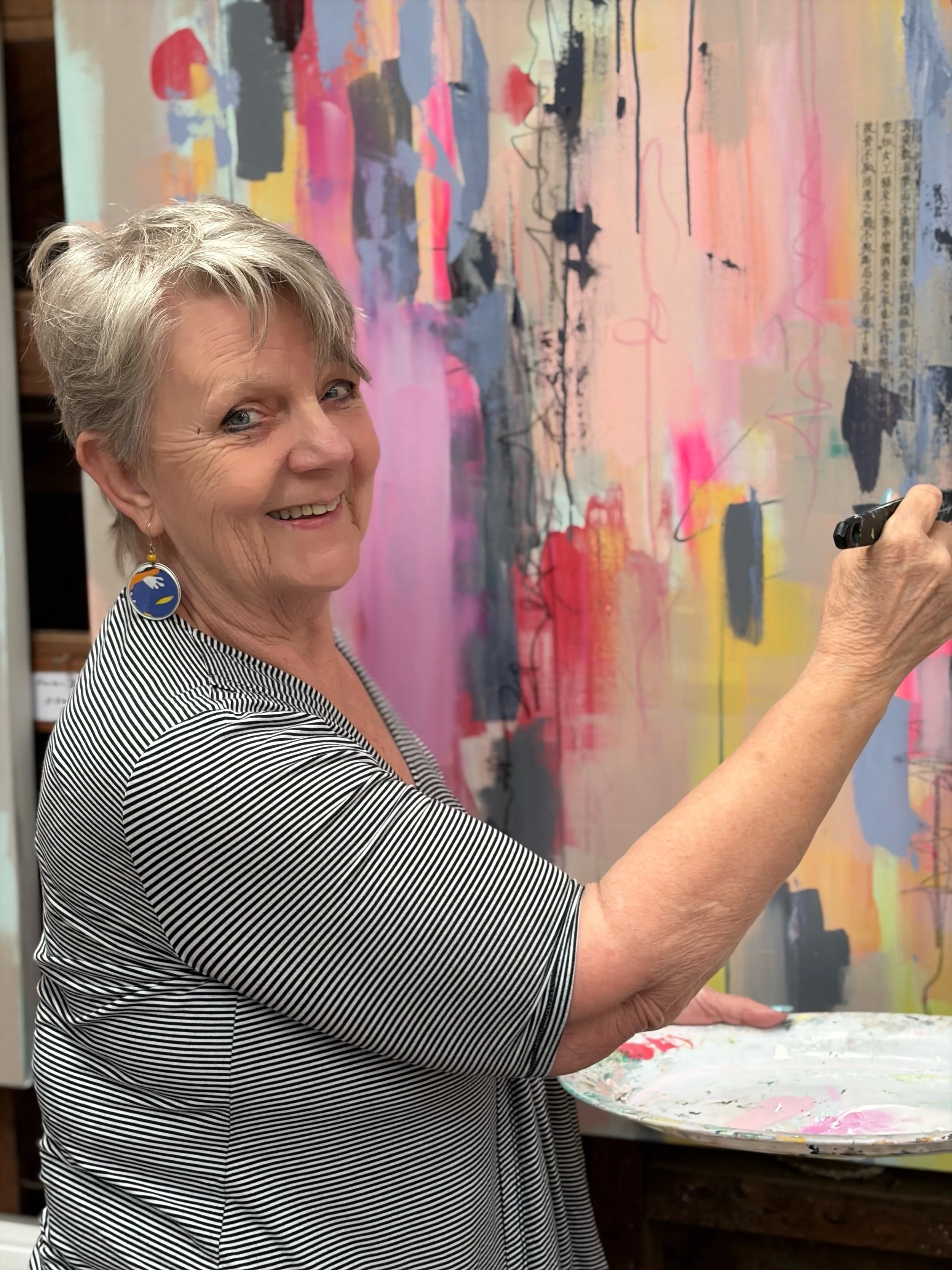A woman with short gray hair, wearing a striped shirt and decorative earrings, smiling while holding a paint palette with pink and other colors in front of an abstract colorful painting.
