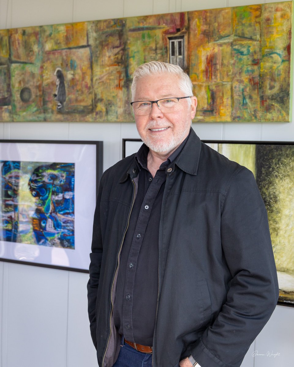 An older man with glasses, white hair, and a beard, smiling, standing in front of colorful artwork at an art gallery.