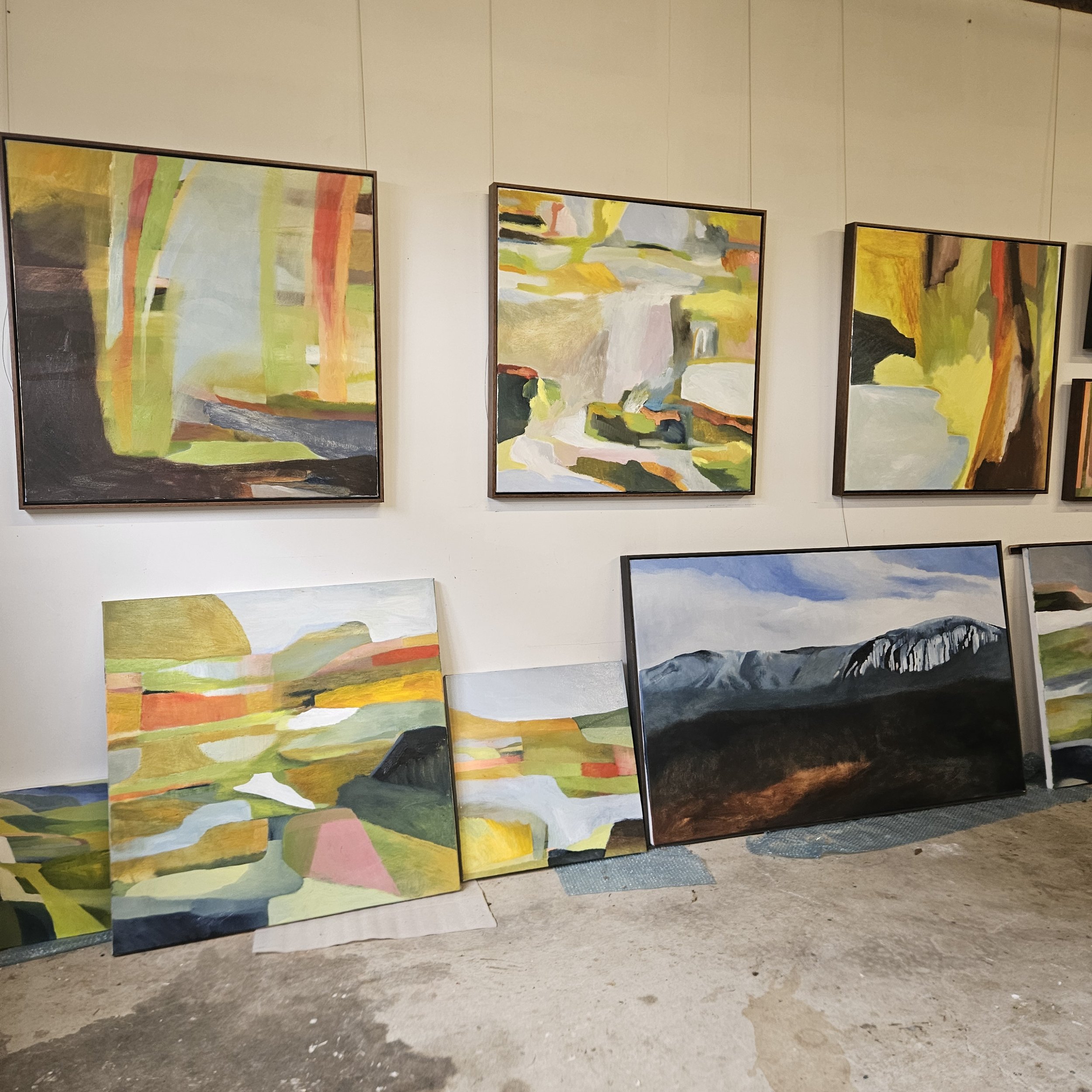 Multiple colorful abstract paintings hanging on a white gallery wall, with some paintings leaning against the wall on the floor.