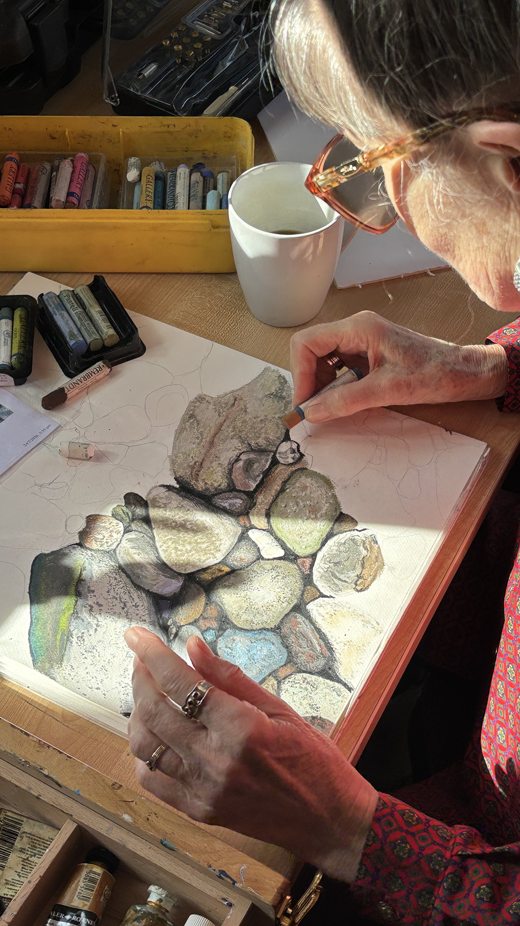 An artist is drawing and coloring a realistic depiction of a pile of stones on a sketchbook. The artist is wearing glasses and a red patterned shirt, working at a desk with art supplies around.