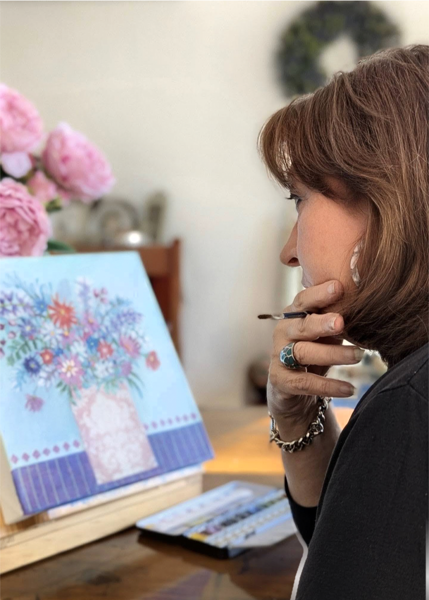 A woman with reddish-brown hair wearing a black top, jewelry, and a ring, is seen from the side, with her hand near her chin, looking at a colorful, floral-patterned artwork or poster on an easel or stand. Pink flowers and a blurred background are also visible.