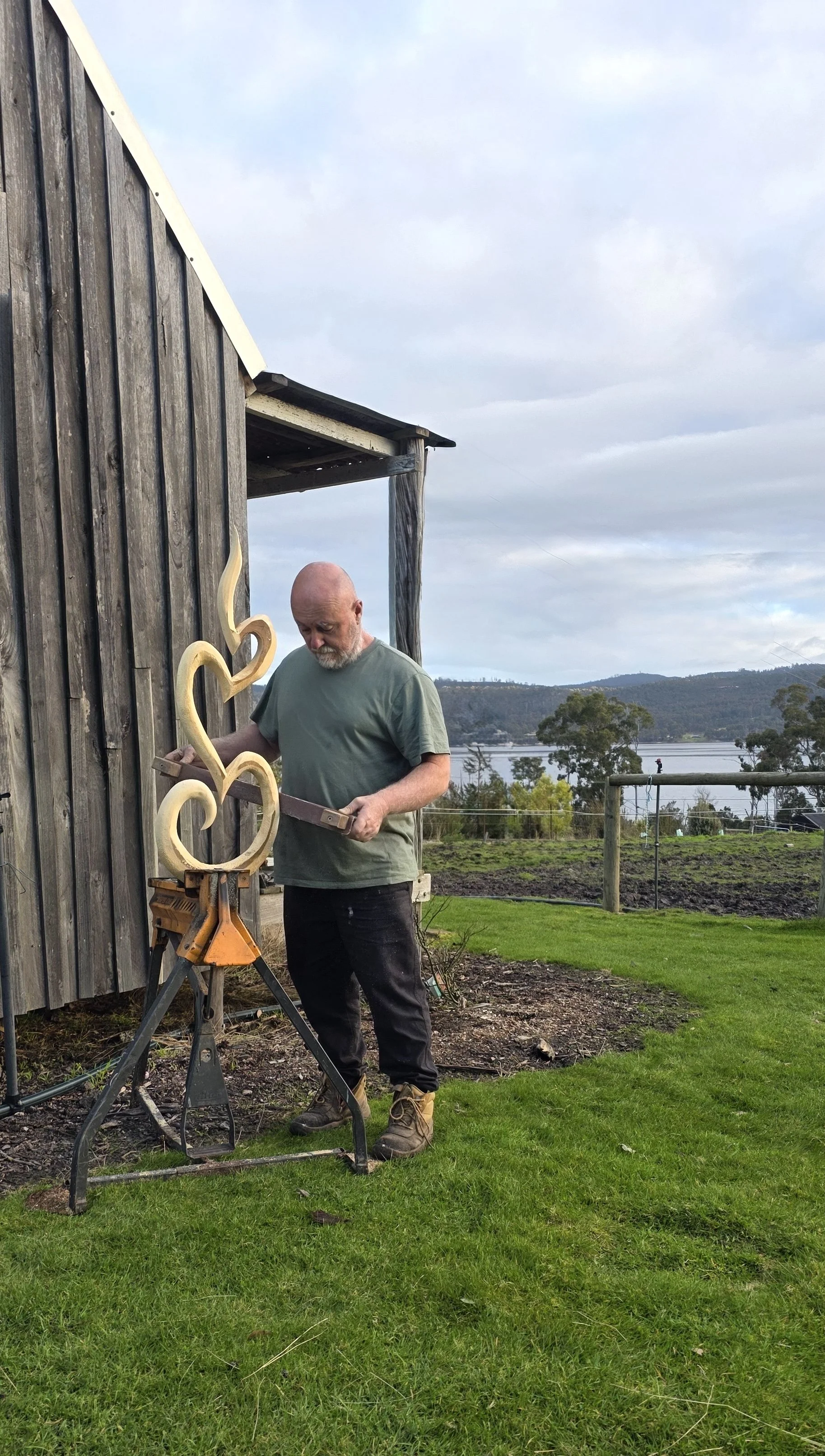 Man working on a metal sculpture outside near a wooden building with a lake and hills in the background.