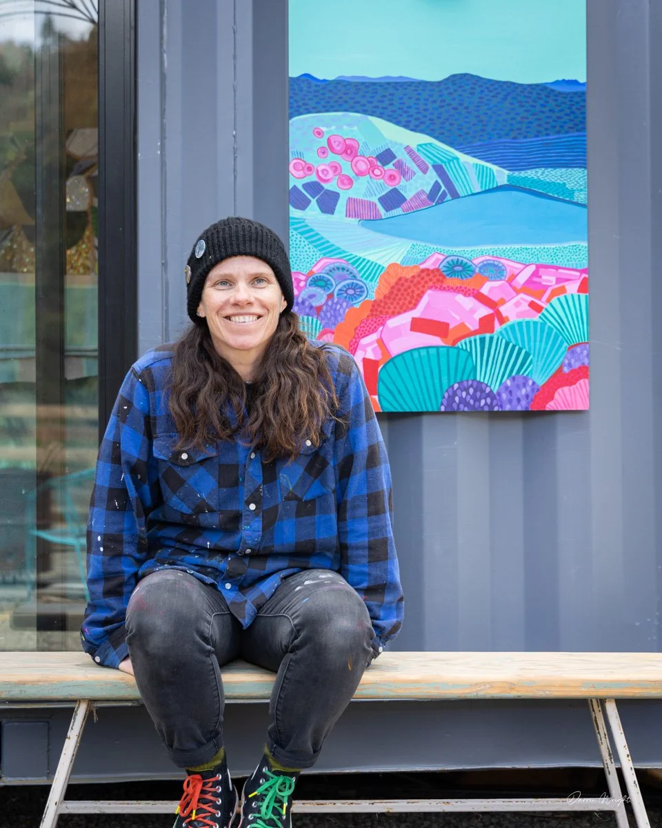 A woman wearing a black beanie, blue plaid shirt, and black pants sitting on a wooden bench in front of a colorful abstract landscape painting.