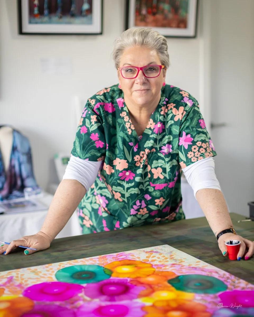 An older woman with short gray hair and pink glasses is leaning over a table, working on a colorful floral artwork that features large, vibrant flowers in pink, purple, yellow, and green. She is wearing a green floral top with pink and peach flowers, and a white long-sleeve shirt underneath. The background shows framed artwork on the wall and an art studio or classroom setting.