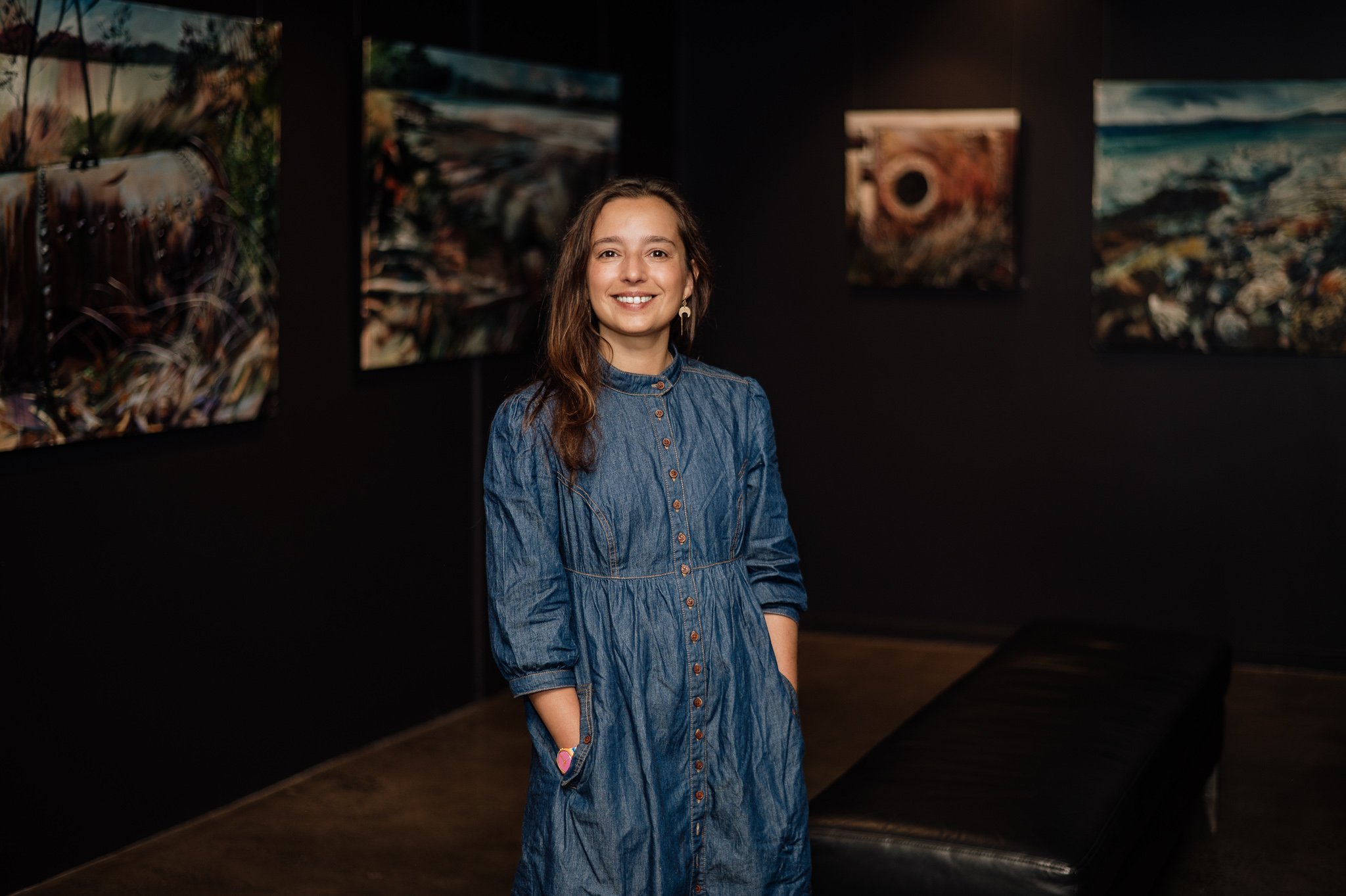 A smiling woman with long brown hair wearing a blue denim dress stands in an art gallery with dark walls, surrounded by several colorful abstract paintings.