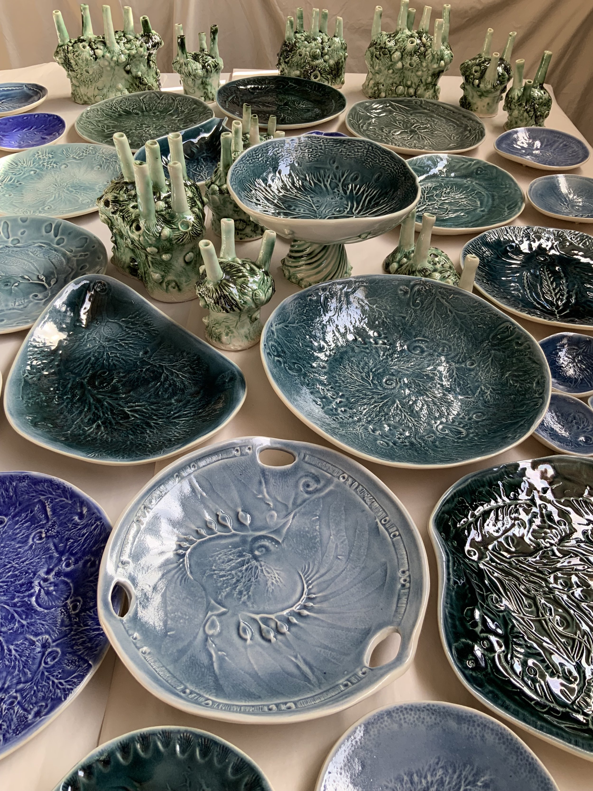 Collection of glazed ceramic plates and bowls with botanical and abstract patterns, some with raised textures, in shades of blue, green, and black.