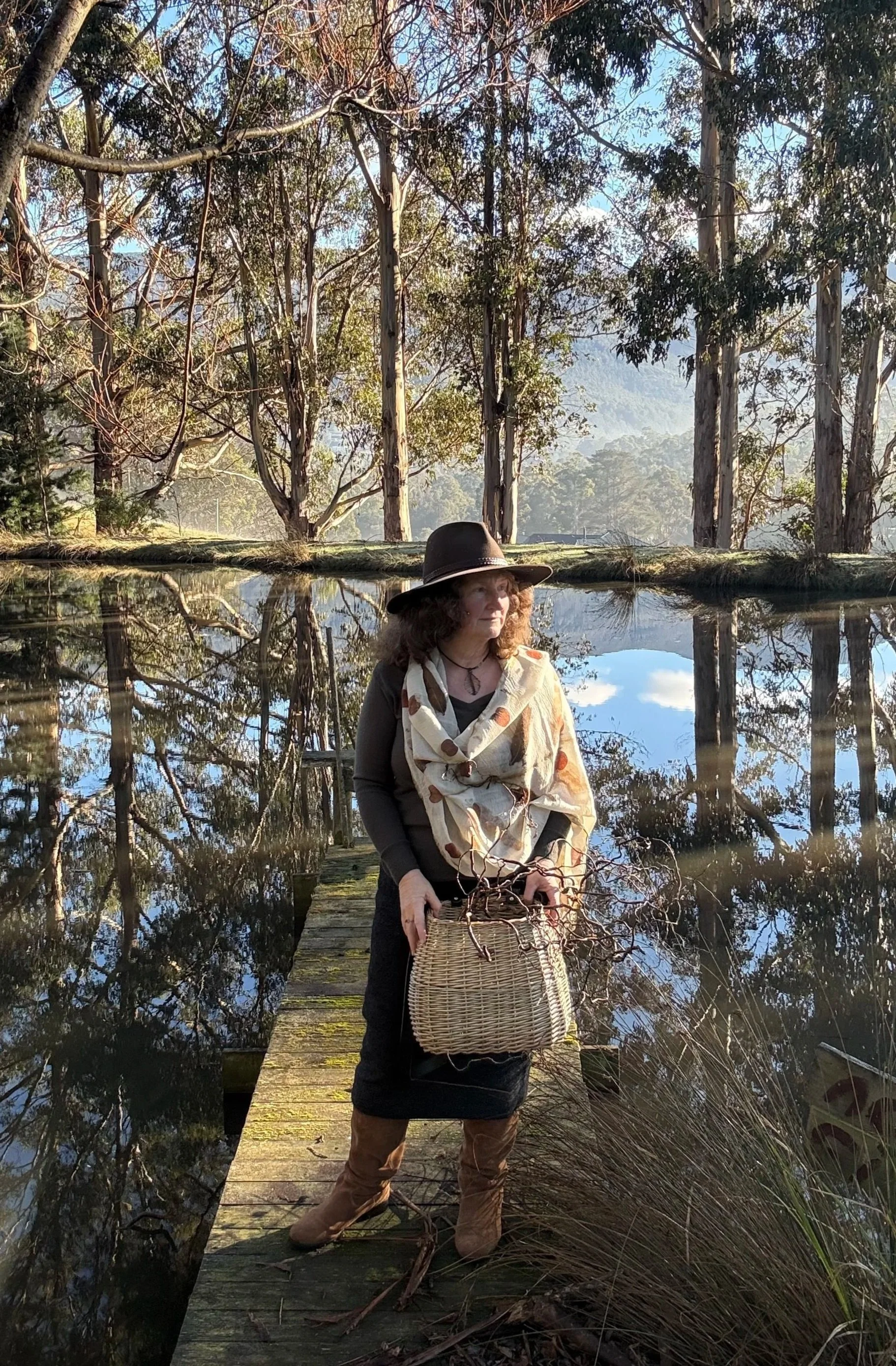 A woman standing on a wooden dock over a calm lake, holding a wicker basket, wearing a large hat and a scarf, with tall trees and mountains in the background during daylight.