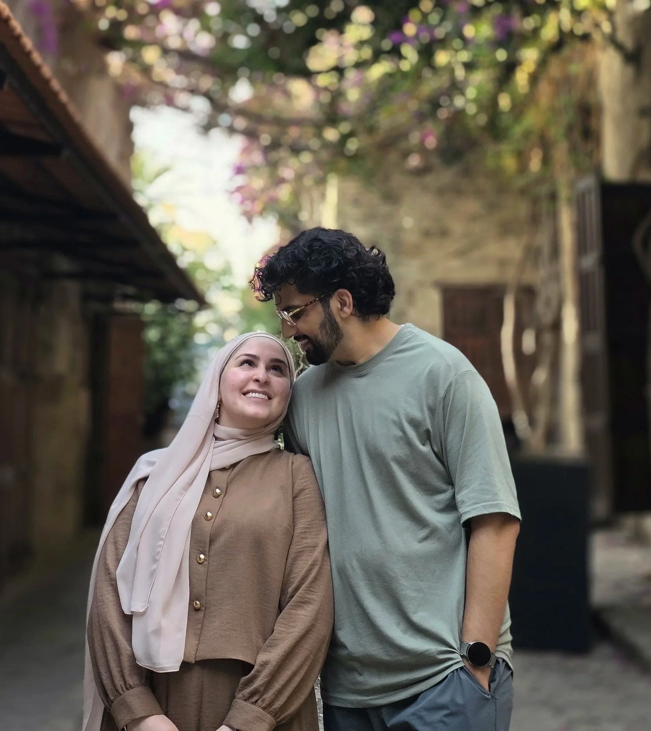 A man with curly black hair, glasses, and a beard stands close to a woman wearing a hijab, smiling and looking at each other. They are outdoors with a blurred background of trees, flowers, and buildings.