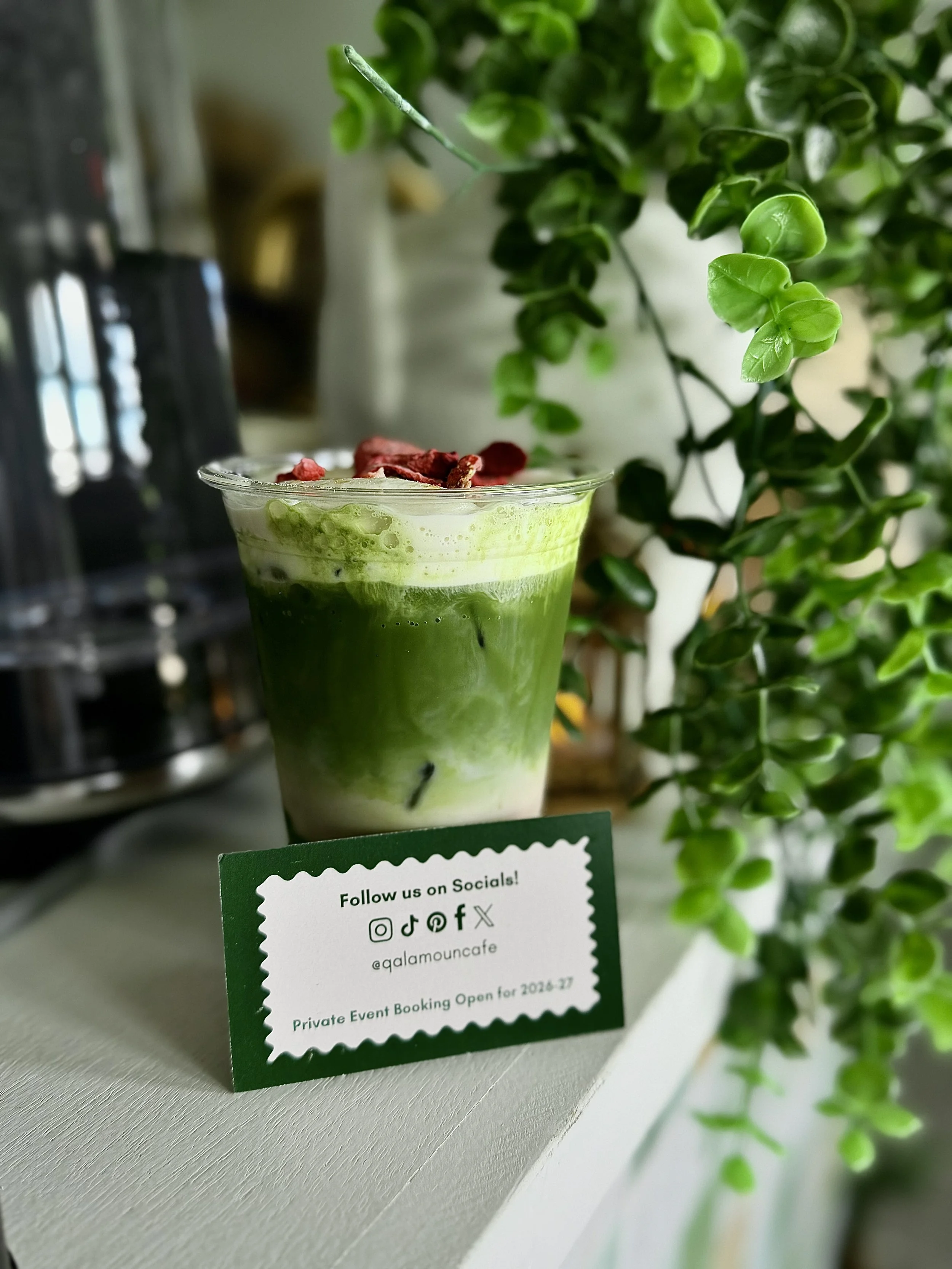 A glass of matcha latte with red berries on top, placed on a white surface near green plants, with a small sign promoting social media and private event bookings.