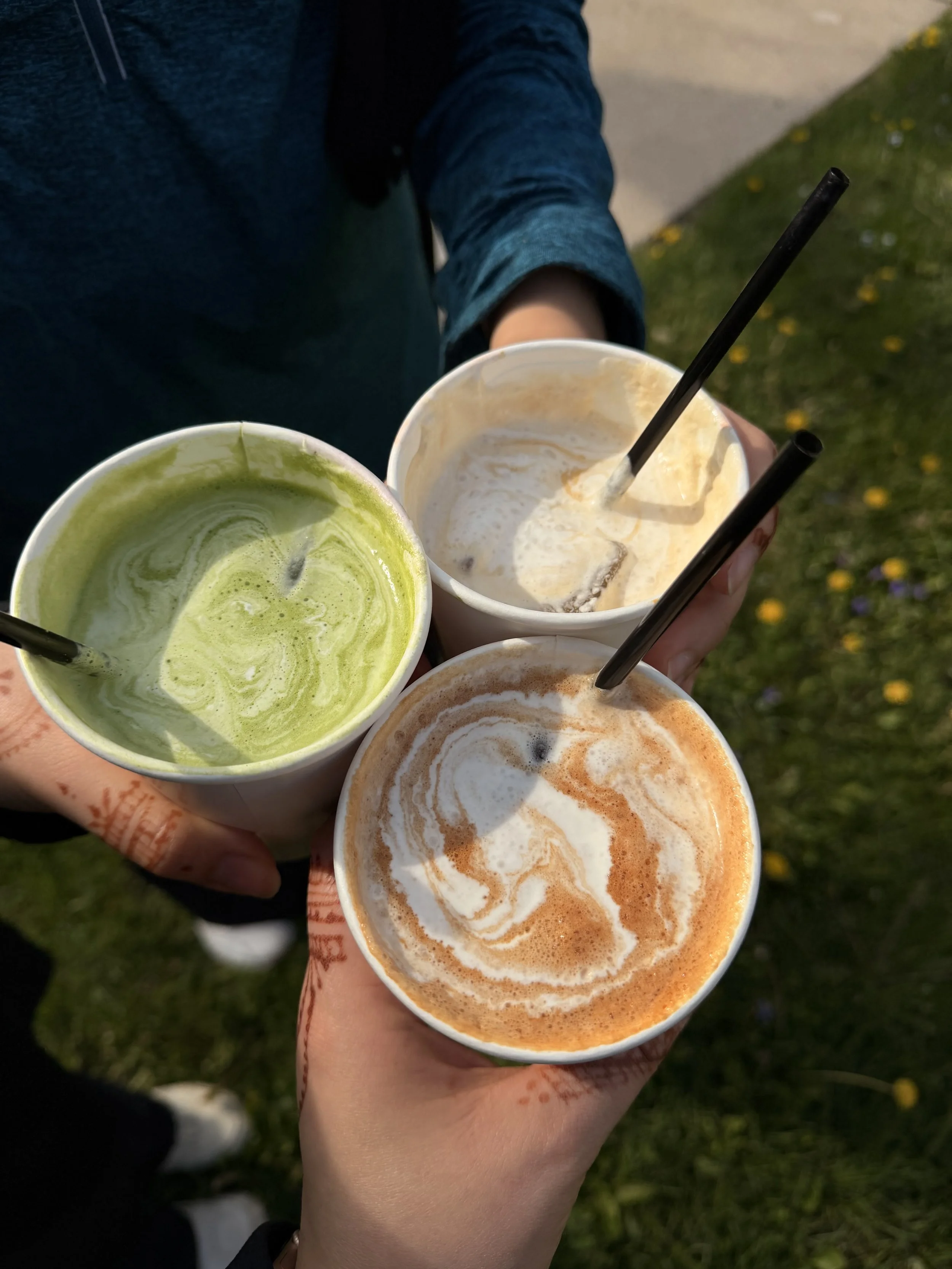 Four cups of colorful drinks held by a person's hand outdoors on a grassy area with small yellow flowers, in the evening or late afternoon.