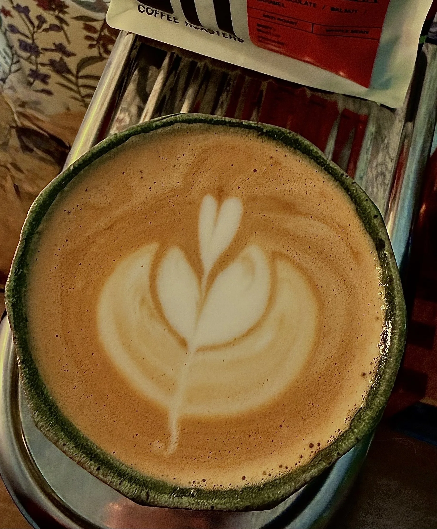 A close-up of a coffee cup with latte art in the shape of a heart, placed on a metal tray. Part of a grocery bag and a wooden surface are visible in the background.
