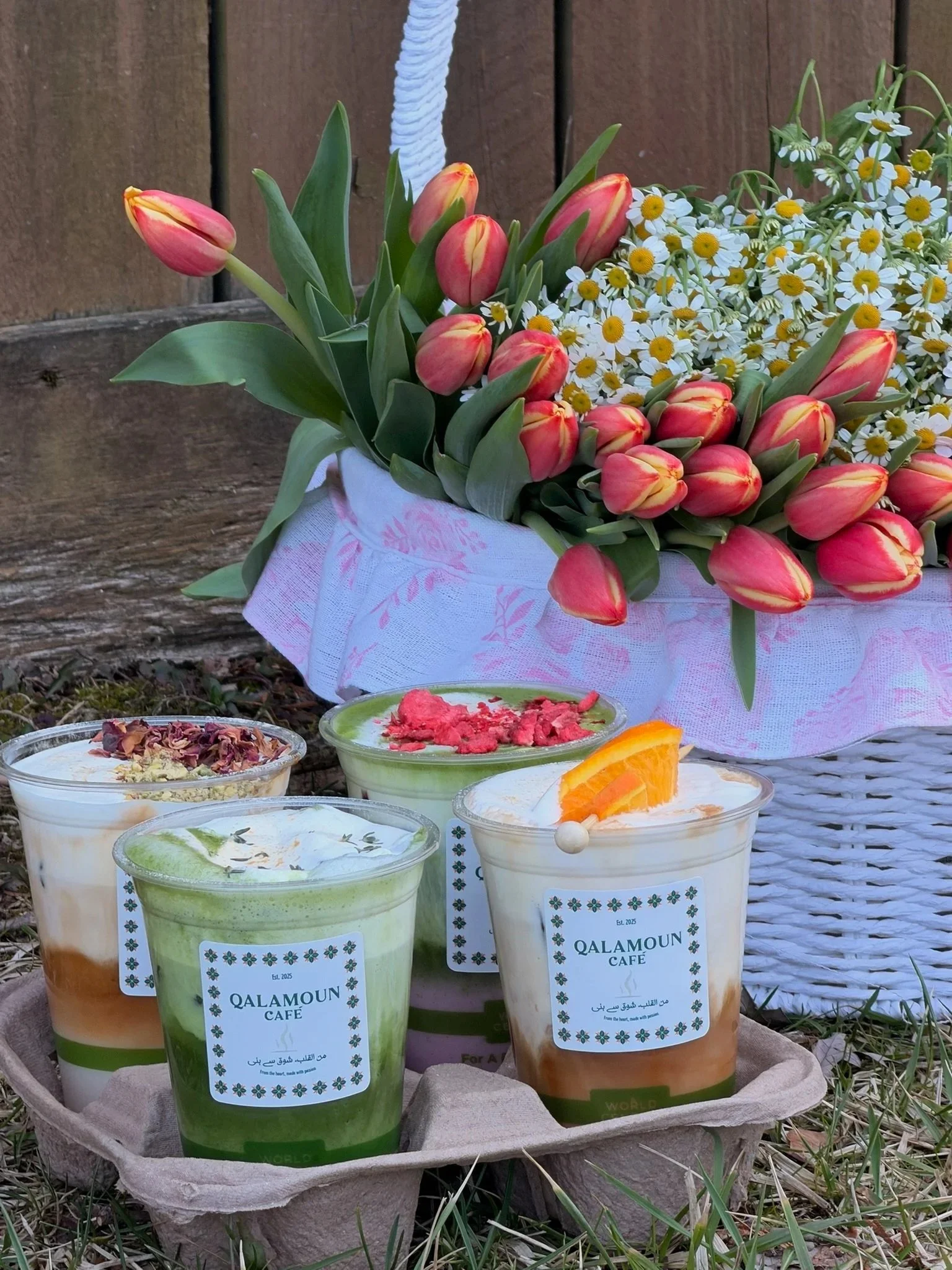 A bouquet of pink and yellow tulips and white daisies in a basket, with several cups of colorful drinks with garnishes and labels that read 'QALAMOUN Café'.