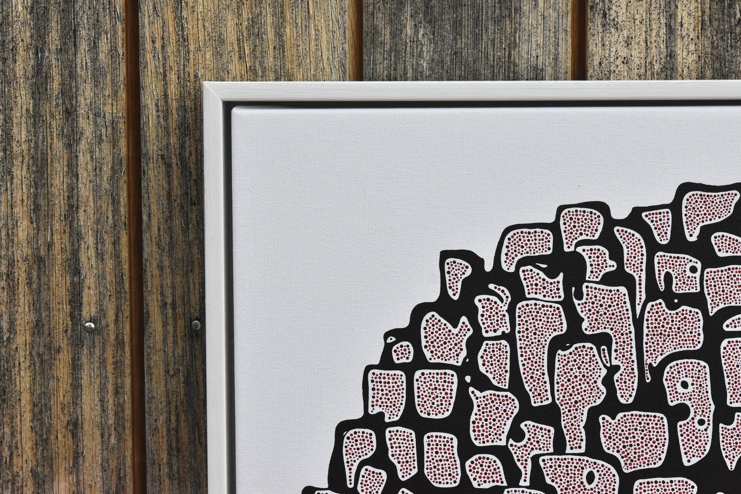 Close-up of a framed piece of artwork with abstract black, white, and red patterns on a white background, leaning against a weathered wooden wall. Julian Oates