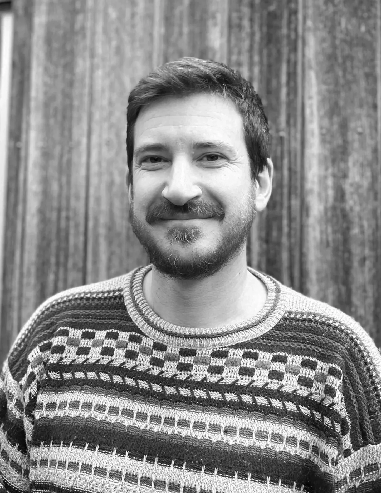 A black-and-white portrait of a smiling man with short hair and a beard wearing a patterned sweater, standing in front of a wooden wall. Julian Oates