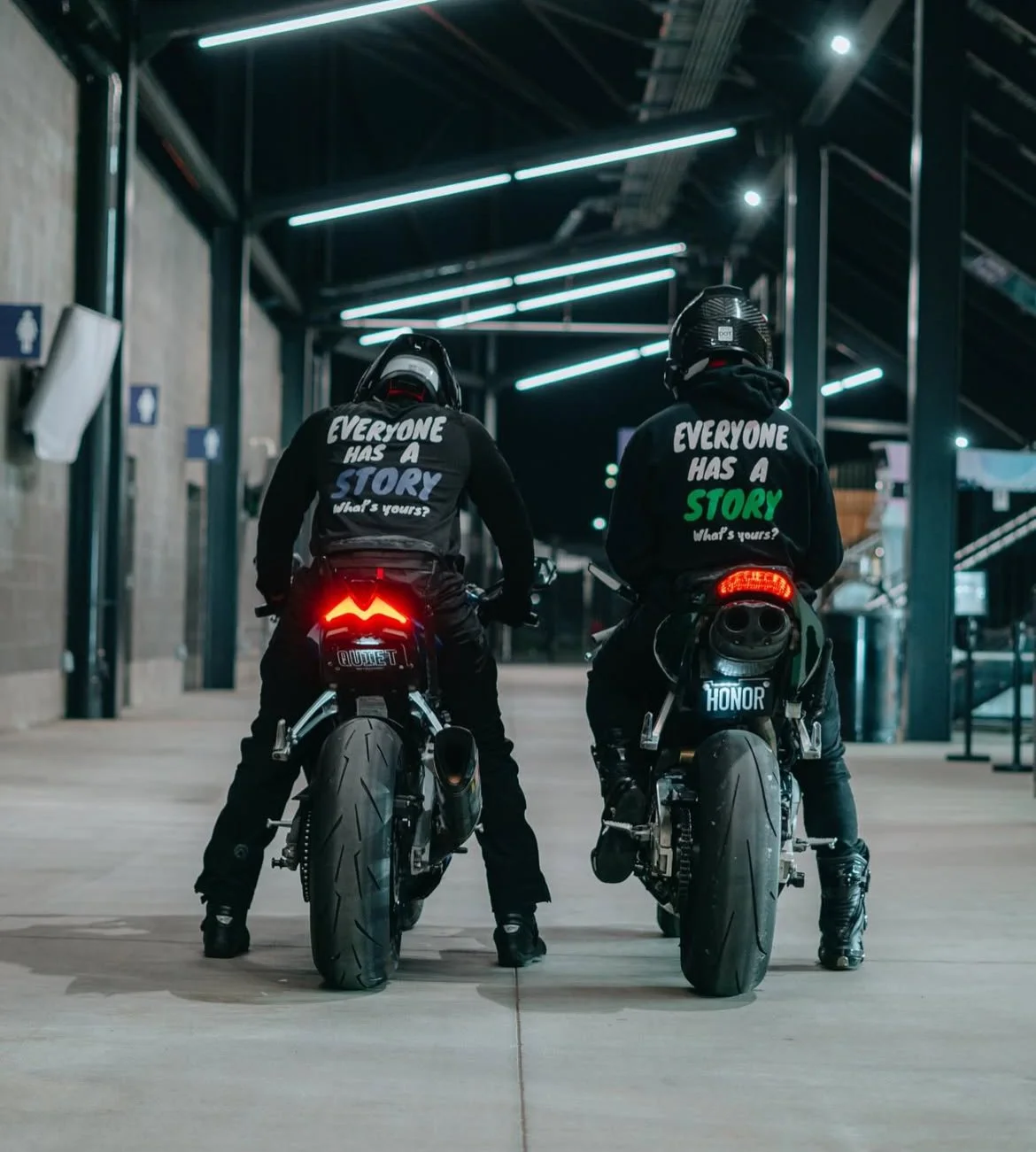 Two motorcyclists in black helmets and jackets sitting on sport bikes in an indoor space. The jackets have quote designs; the left says "Everyone has a story, what's yours?" in blue, and the right says "Everyone has a story, What's yours?" in green. 