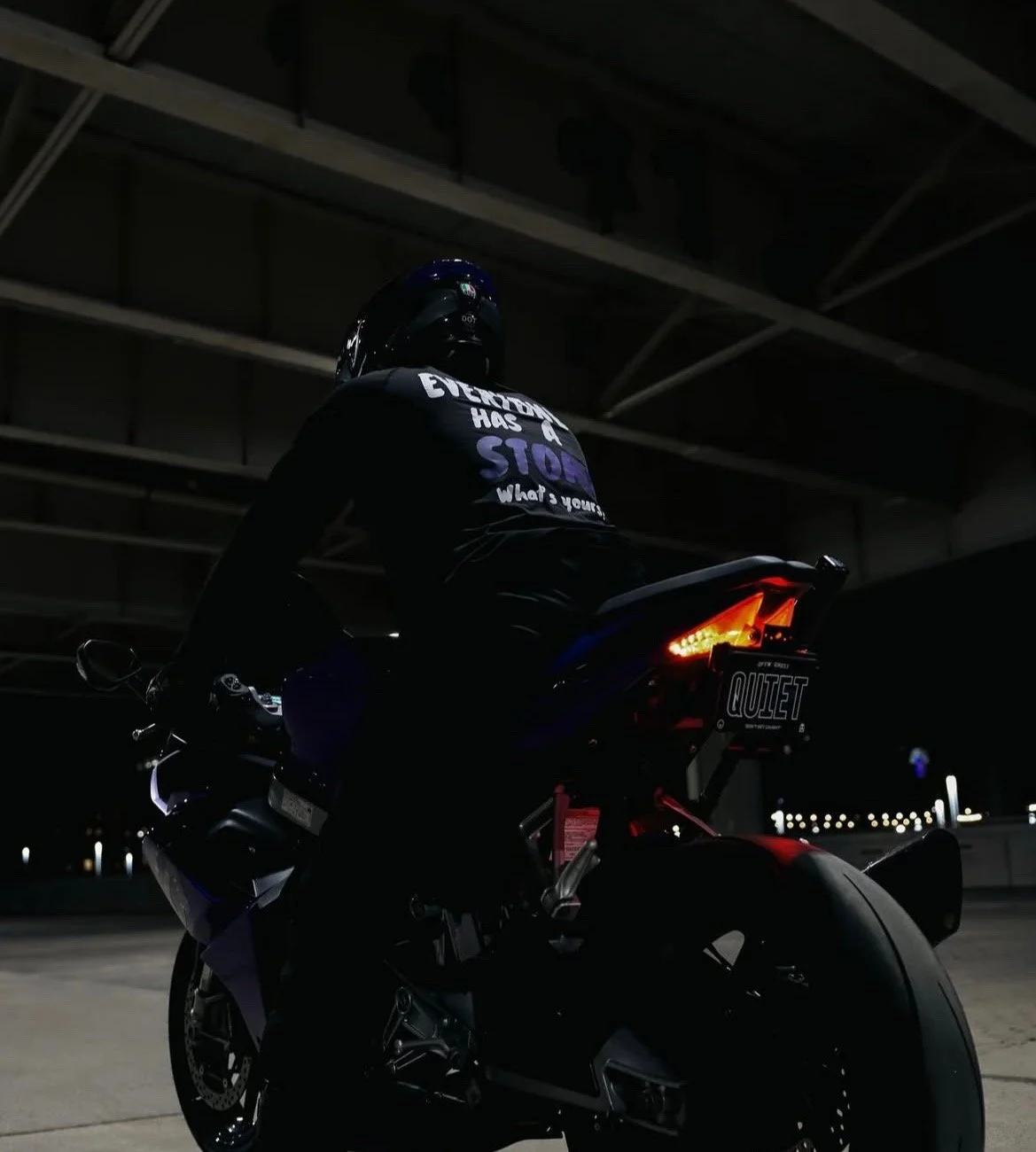 A person in a motorcycle helmet and dark clothing sitting on a black sports motorcycle at night with illuminated taillights and city lights in the background.