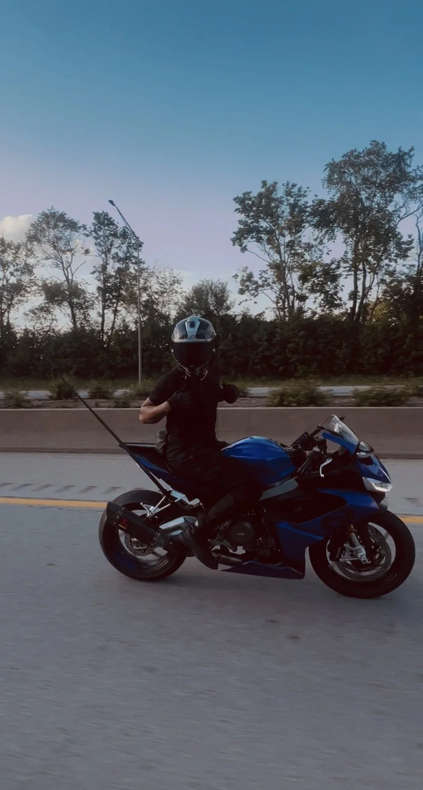 A person wearing a black helmet and black shirt riding a blue motorcycle on a road with trees and a streetlamp in the background during dusk.
