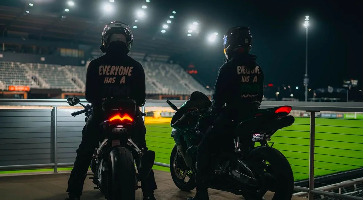 Two motorcyclists with helmets sitting on motorcycles at a stadium at night, overlooking a lit sports field and empty bleachers.