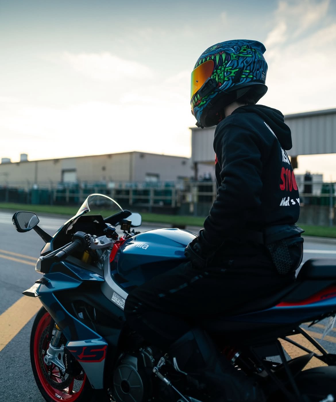 A person sitting on a blue and black motorcycle, wearing a colorful helmet with a tinted visor and a black hoodie with red and white text, in an industrial area during daylight.