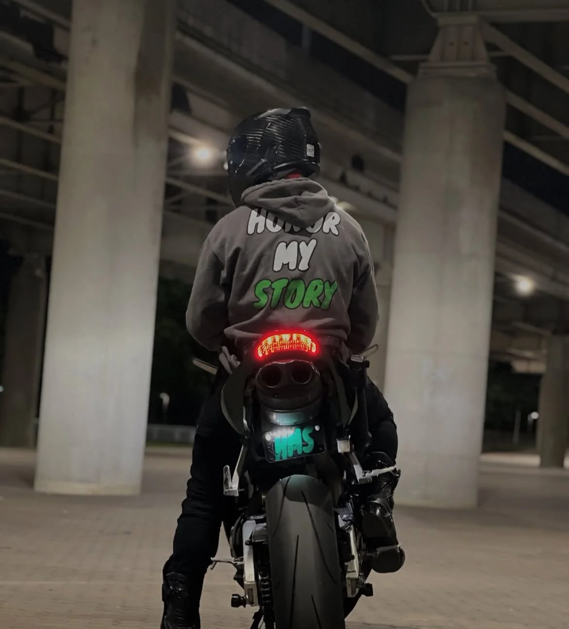 A person wearing a motorcycle helmet and a hoodie that says 'HONOR MY STORY' is sitting on a black motorcycle, illuminated by a red rear light, under a bridge at night.