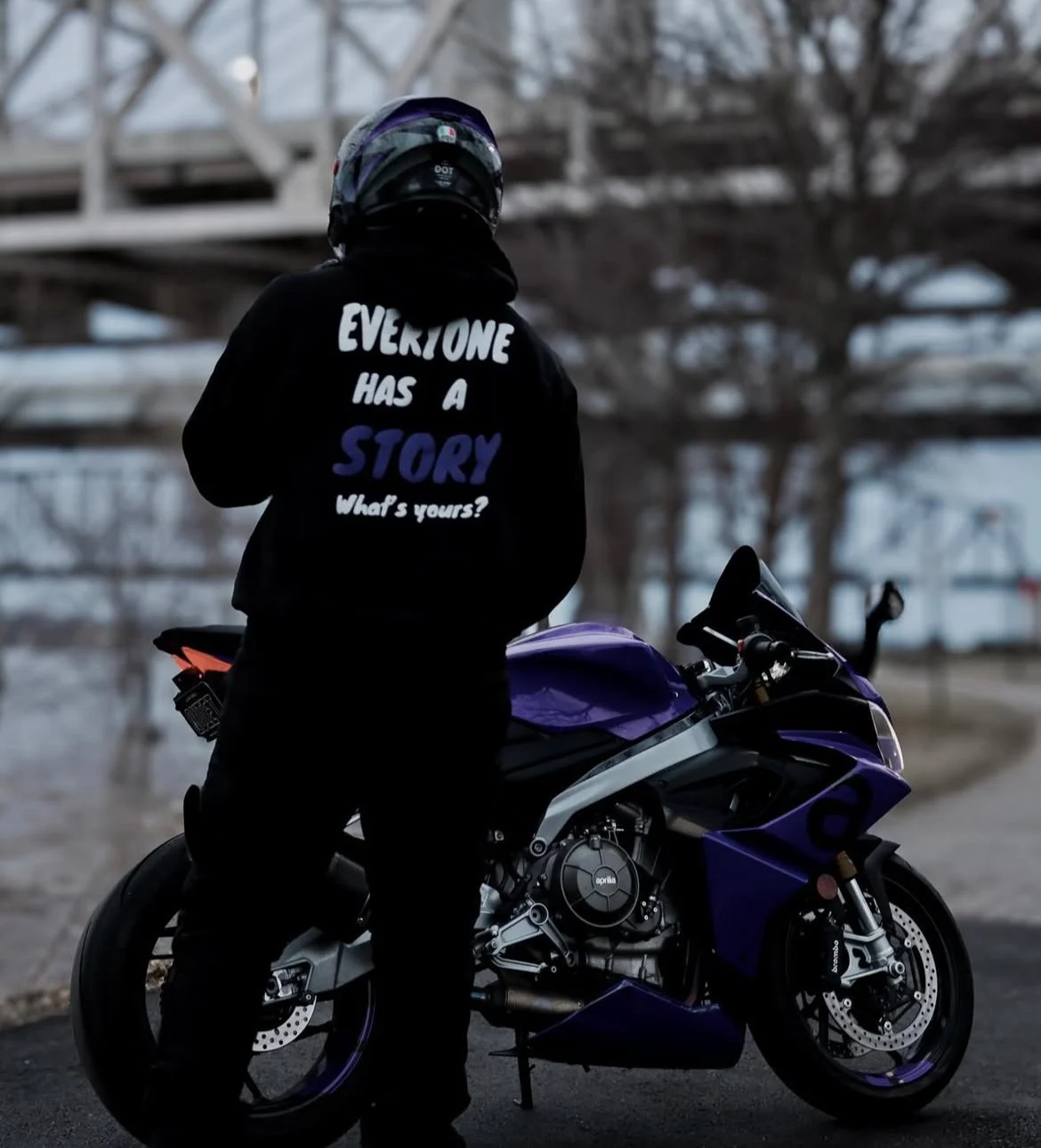 Person wearing a helmet and hoodie standing next to a purple motorcycle under an overpass.