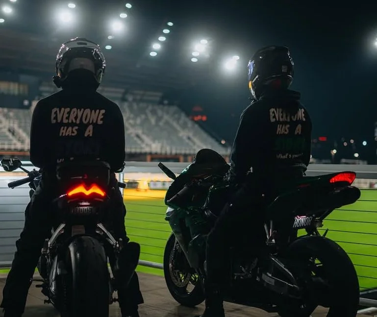 Two motorcyclists seated on their bikes at a racetrack at night, wearing jackets with the phrase "Everyone has a" on the back, overlooking the track illuminated by bright lights.