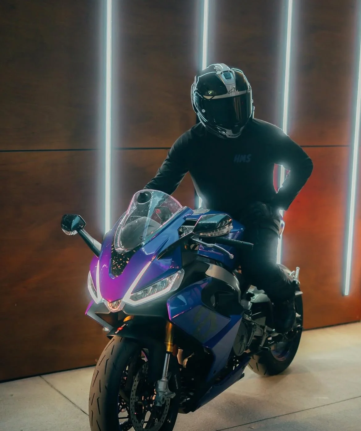 Person wearing a black helmet and black jacket sitting on a blue sports motorcycle inside, with vertical light strips on a wooden wall in the background.