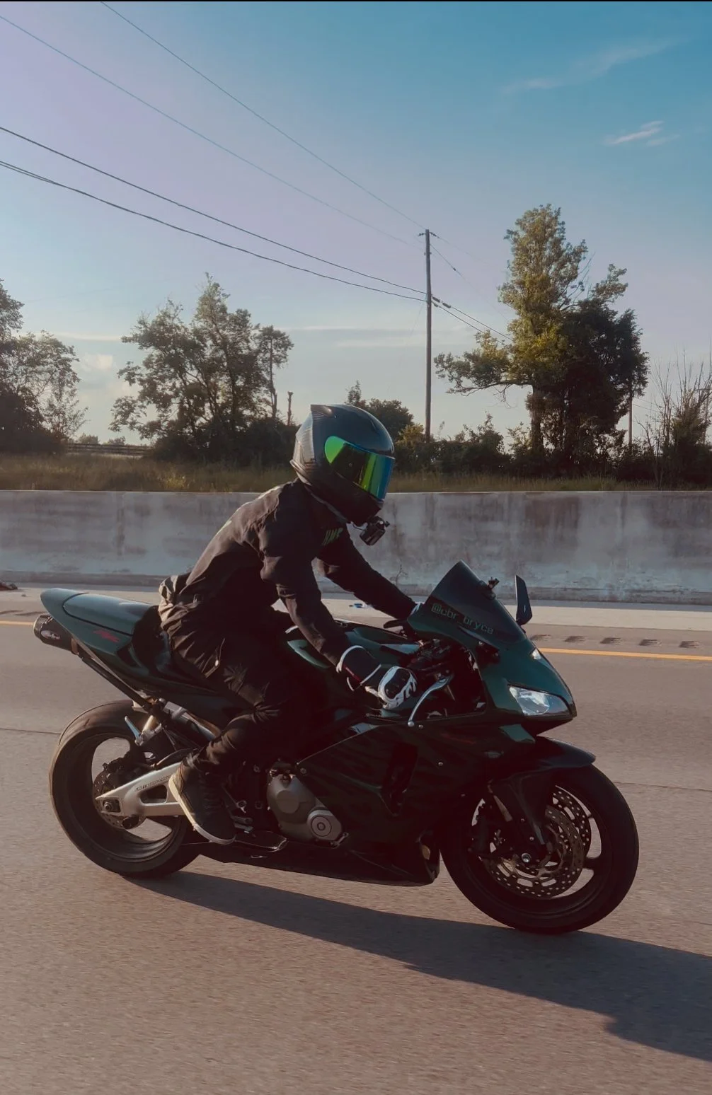A person wearing a black jacket, black pants, white gloves, and a black helmet with reflective visor riding a black sports motorcycle on a highway during the day.