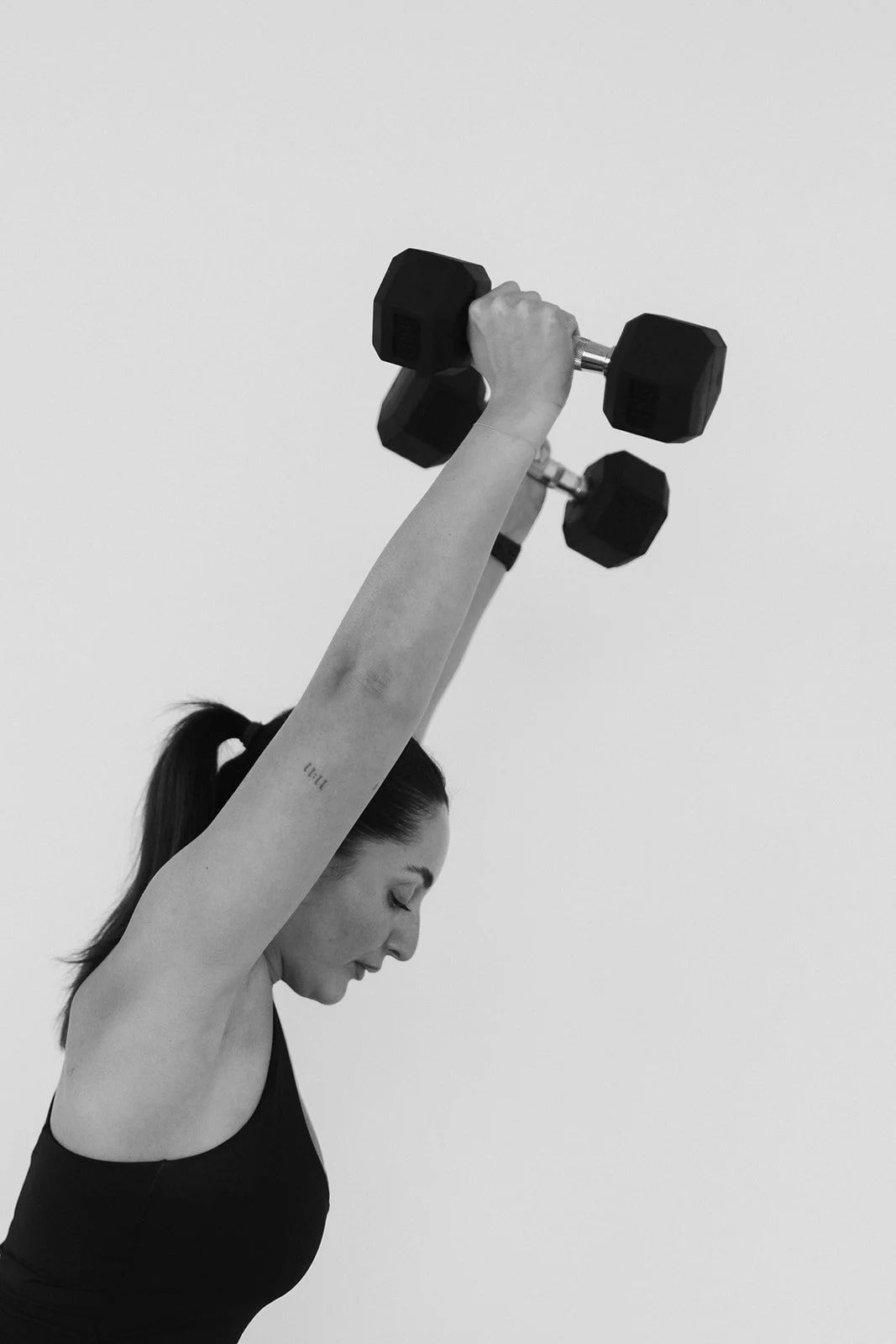 A Hot Mess Instructor lifting a dumbbell with one arm, head tilted down, against a plain background.