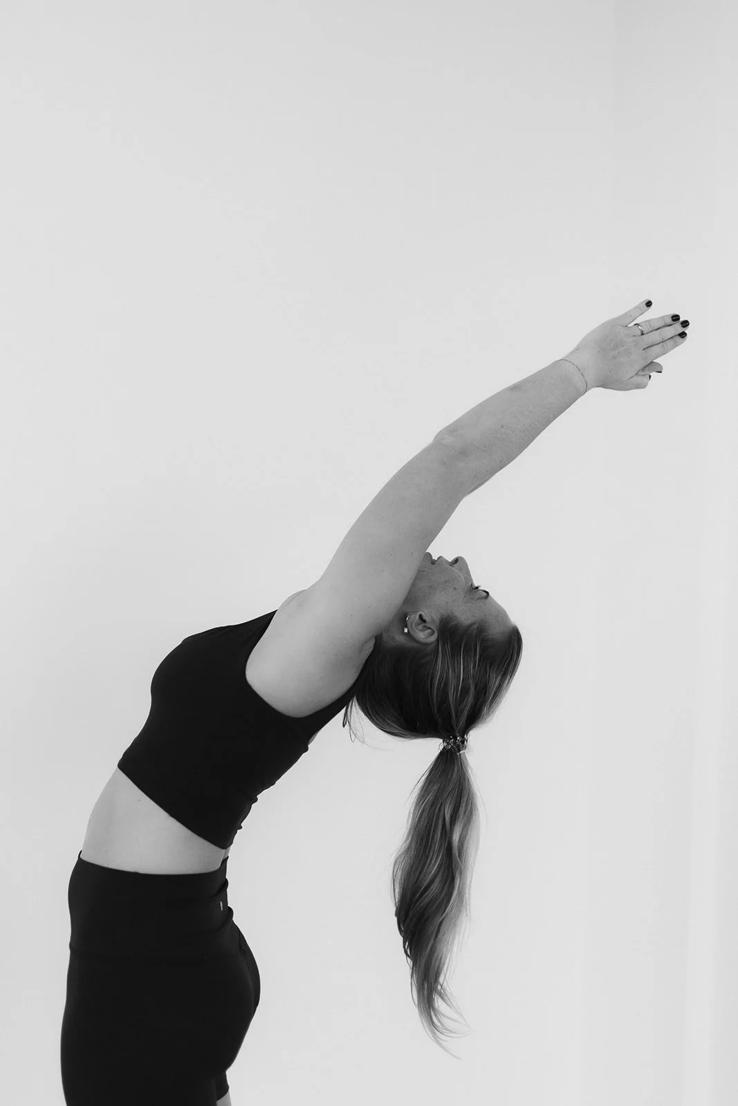 A Hot Mess Instructor performing a yoga pose in a studio with a plain background, wearing a black sports bra and matching high-waisted leggings.