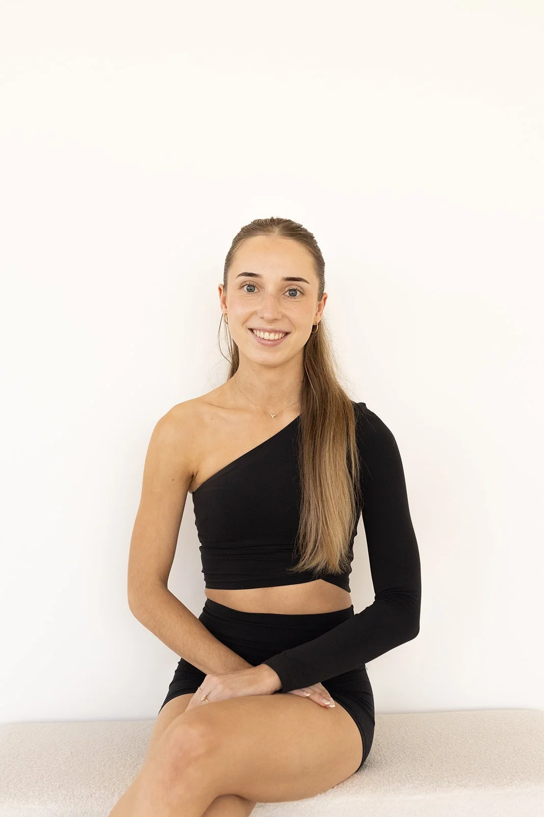A Hot Mess Instructor with long light brown hair in a black, one-shoulder crop top and matching shorts, sitting on a beige bench against a plain white wall, smiling at the camera.