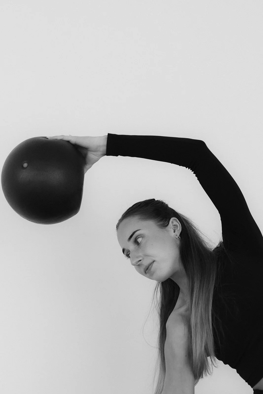 A Hot Mess Instructor performing a pilates pose with a black pilates ball held above her head, against a plain white background.