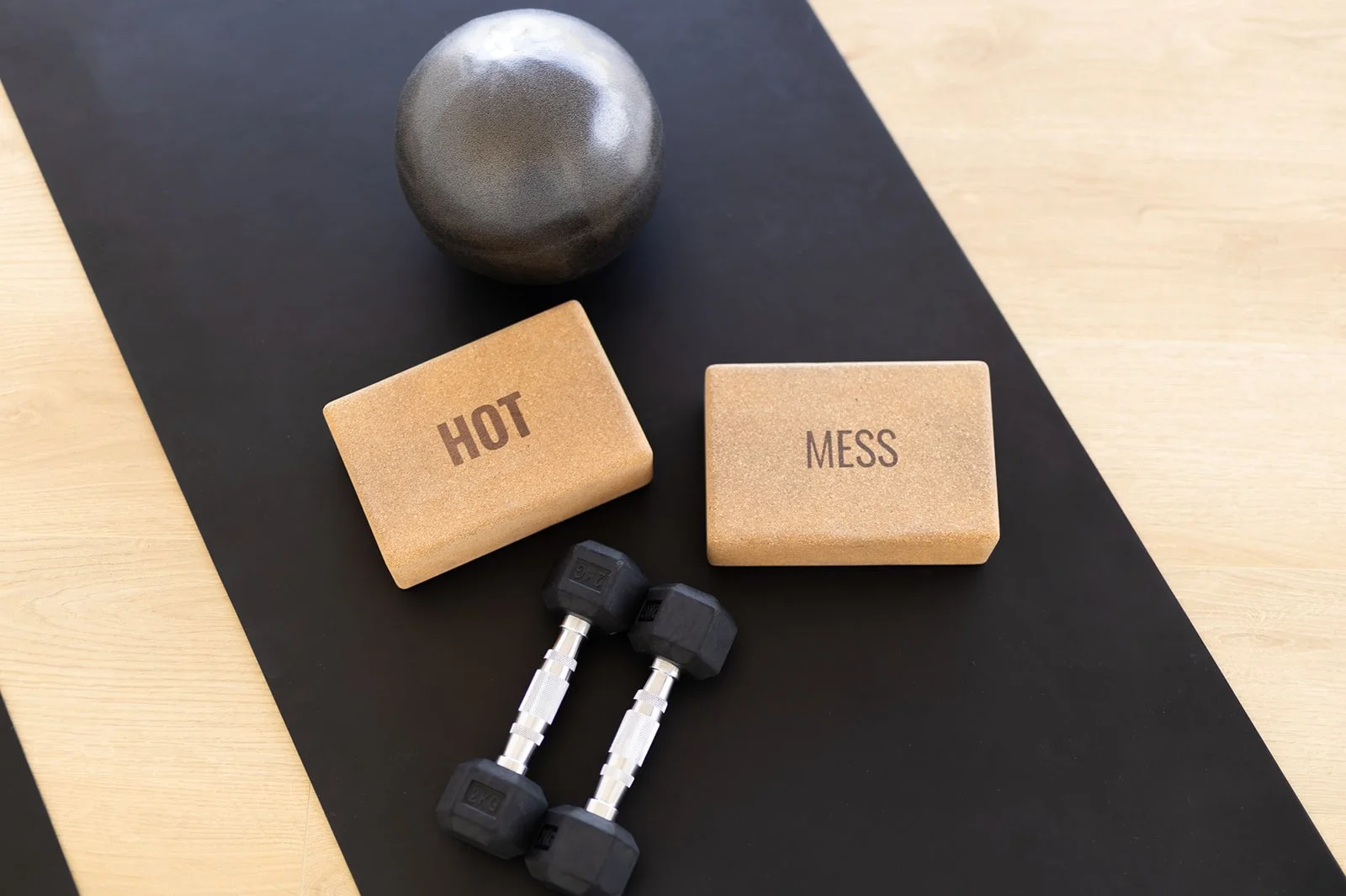 Workout equipment including a black kettlebell, two beige weight blocks labeled 'HOT' and 'MESS', and two black dumbbells on a black workout mat on a light wooden floor.