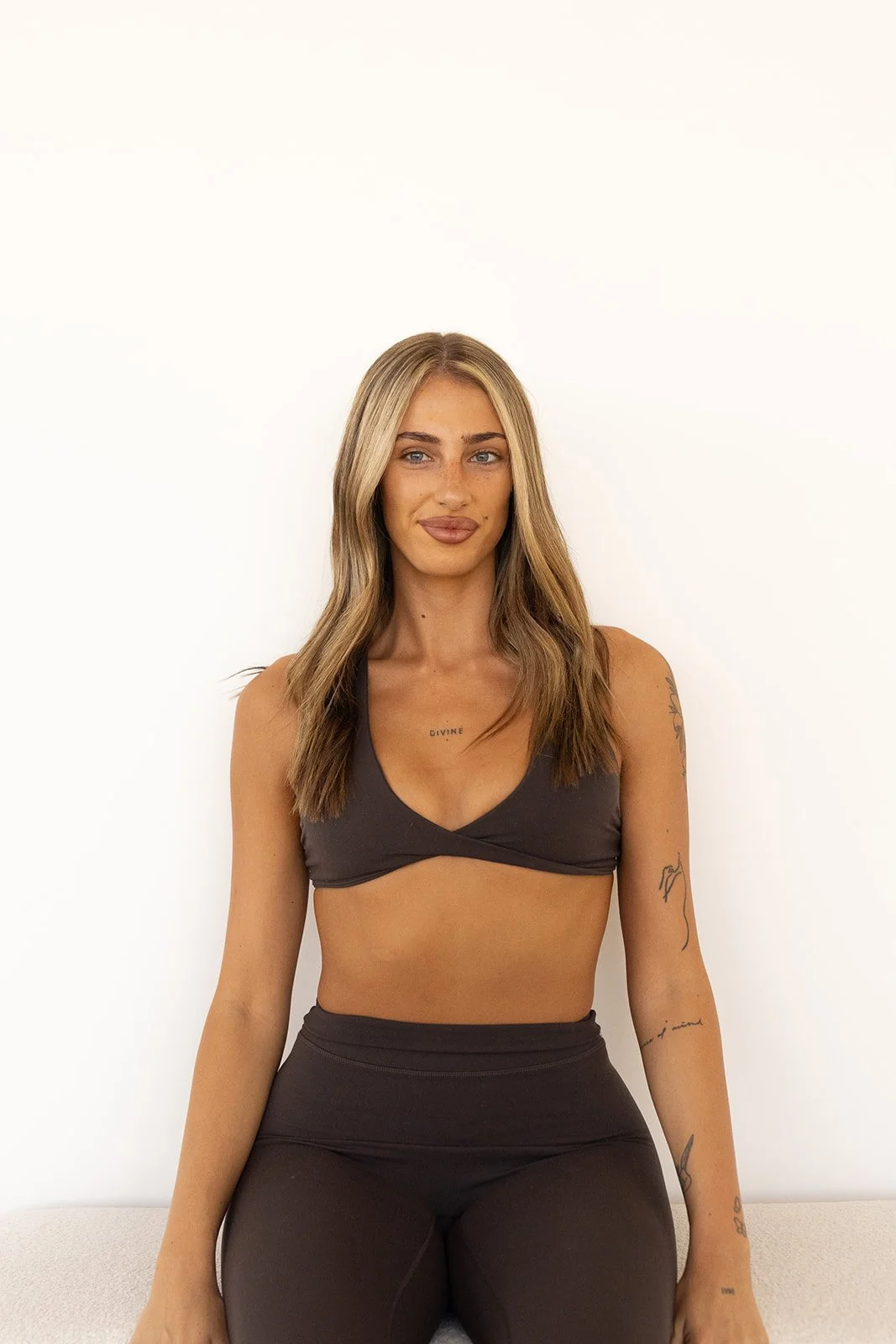 A Hot Mess instructor with long blonde hair, wearing a dark sports bra and black leggings, sitting against a plain white background.