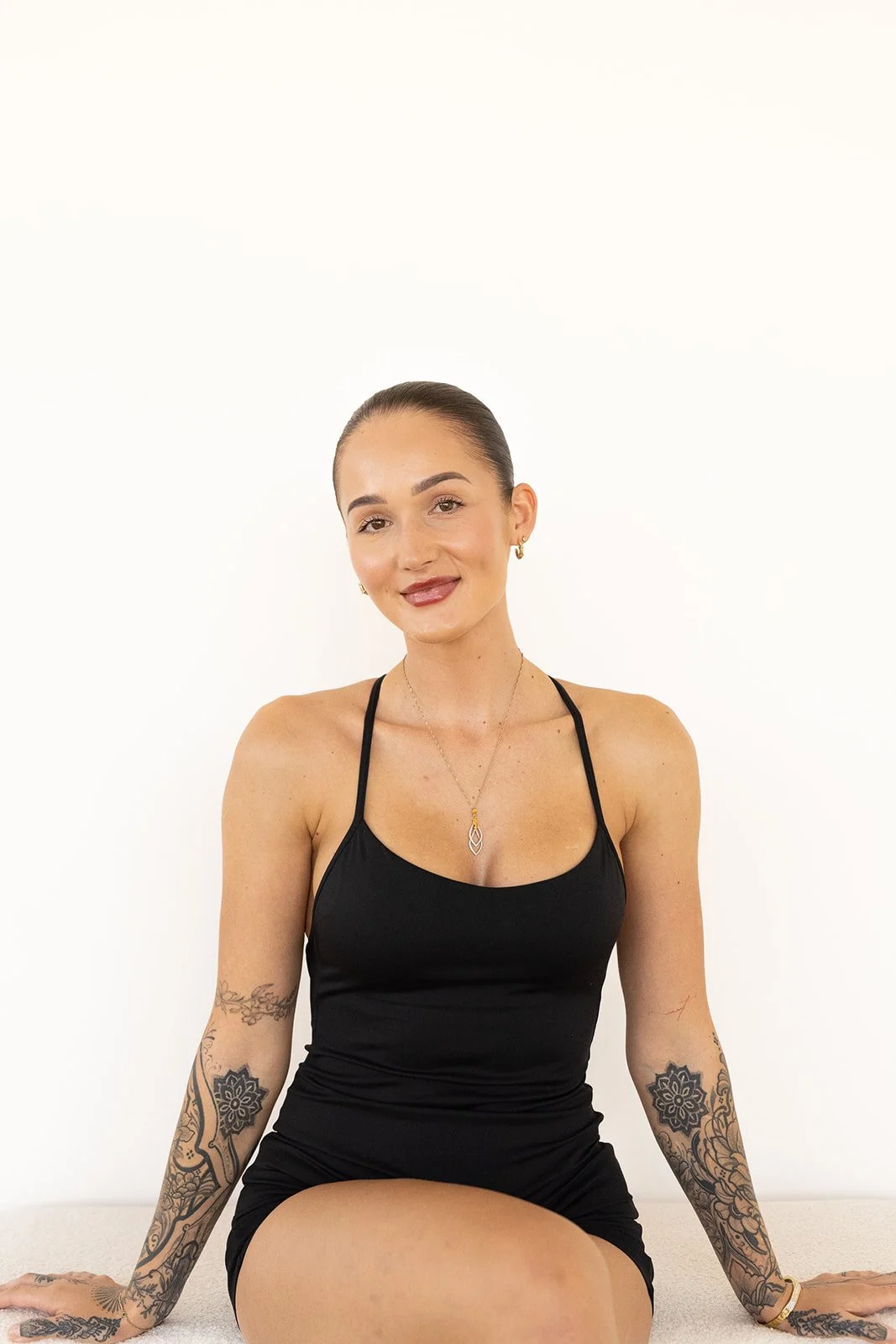 A Hot Mess Instructor with tattoos on arms, wearing black dress, smiling, sitting on white surface against white background.