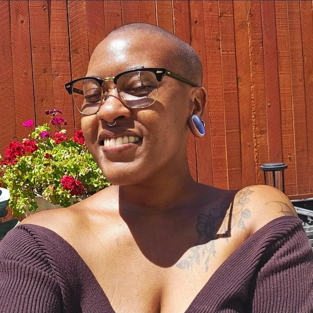 A person with short hair, glasses, and earrings smiling outdoors with a wooden fence and colorful flowers in the background.