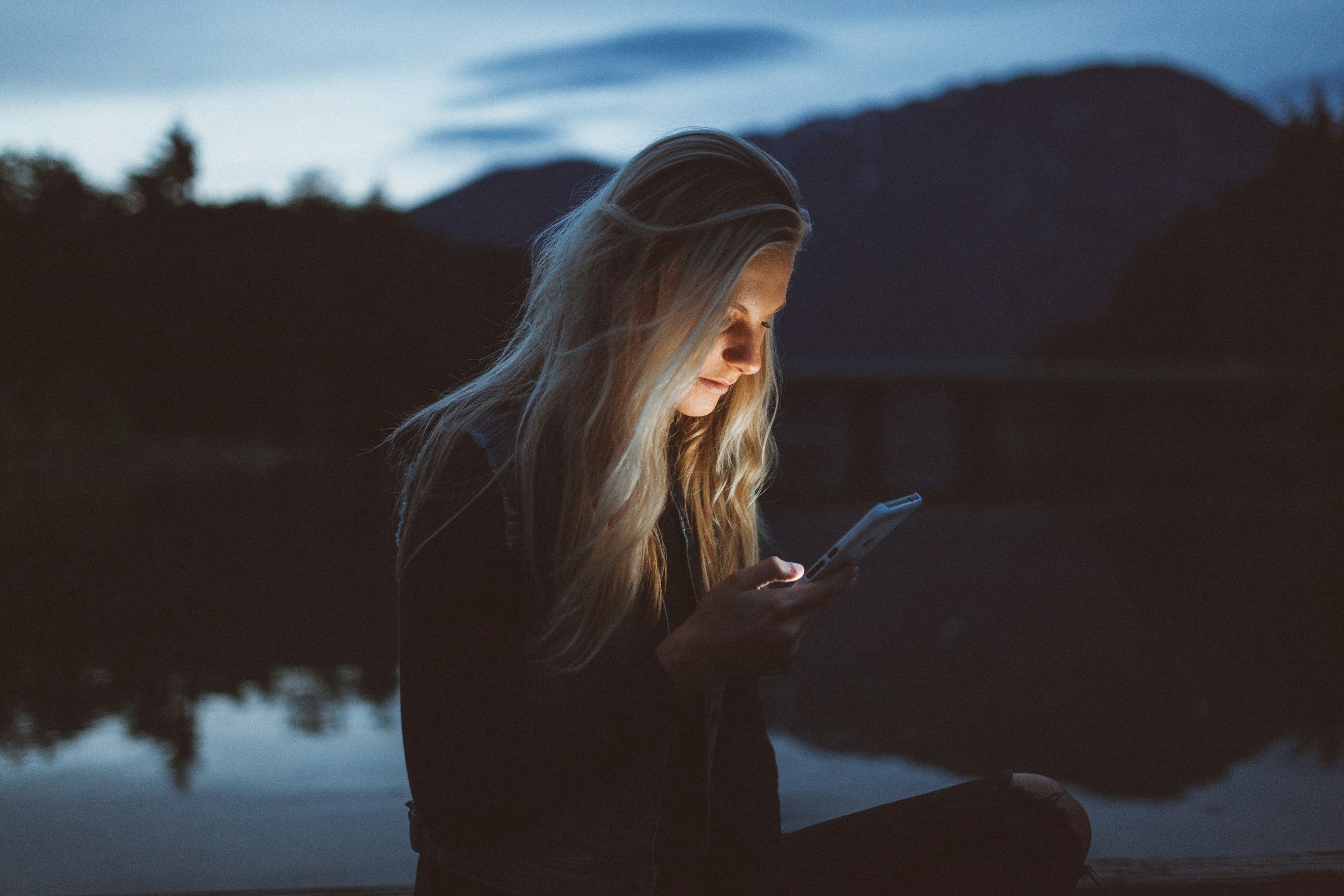 A young woman with long blonde hair looking at her phone during twilight outdoors near a body of water and mountains.