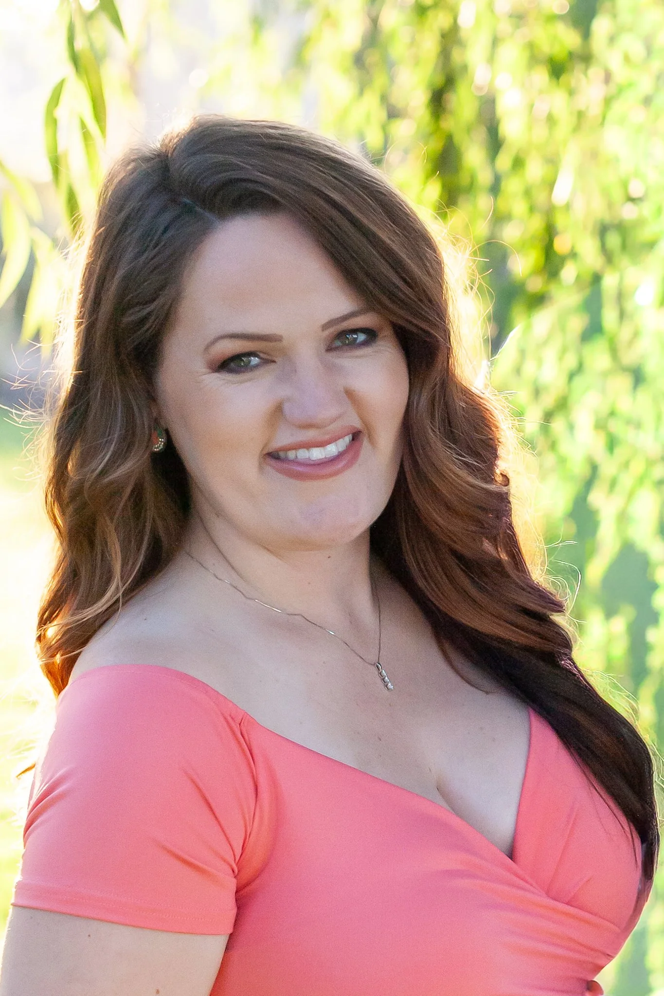 A woman with long wavy brown hair wearing a coral dress with a V-neckline, smiling outdoors with greenery in the background.