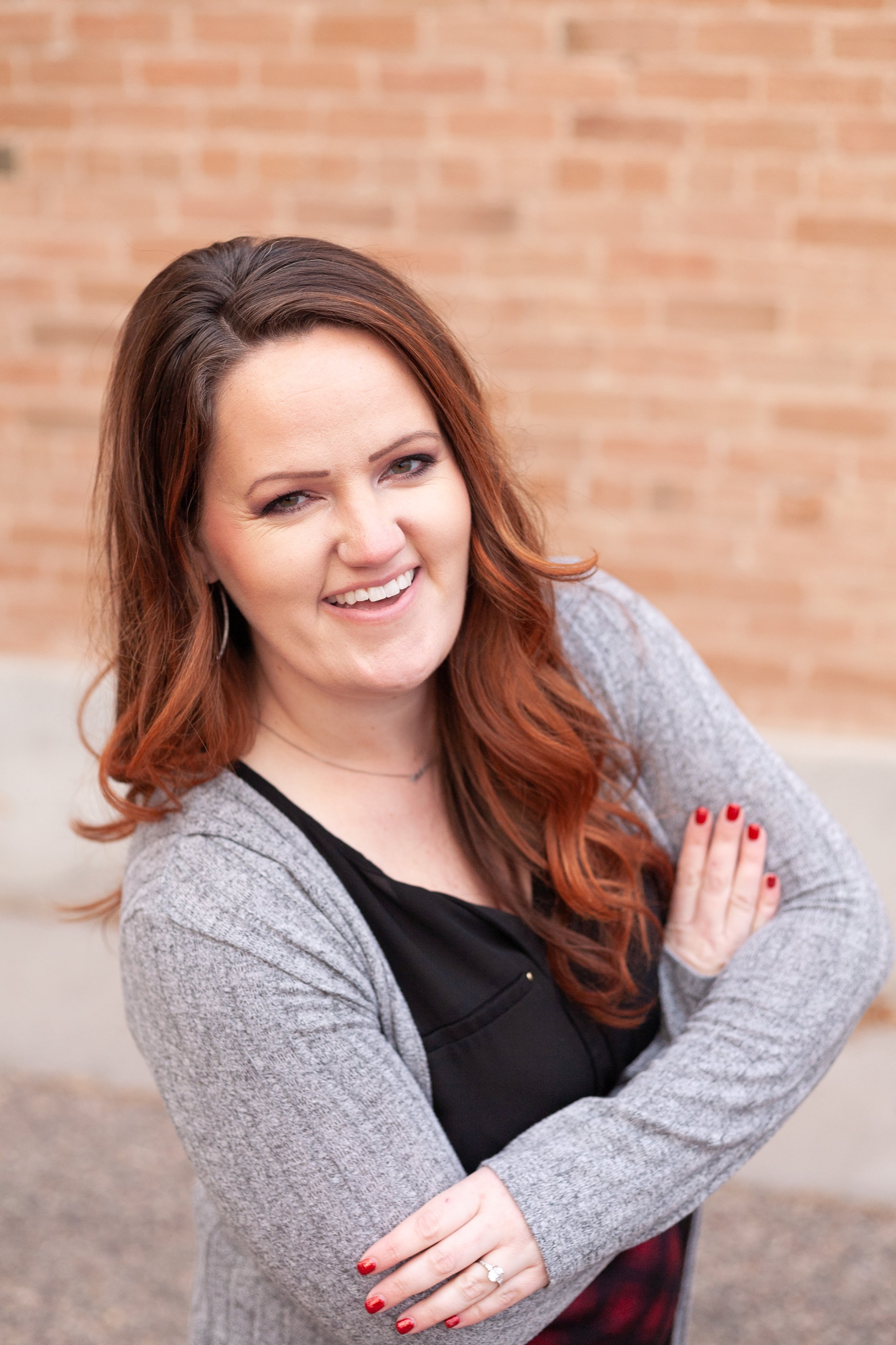 A woman with long reddish-brown hair, smiling and crossing her arms, wearing a black top and gray cardigan, standing in front of a brick wall.