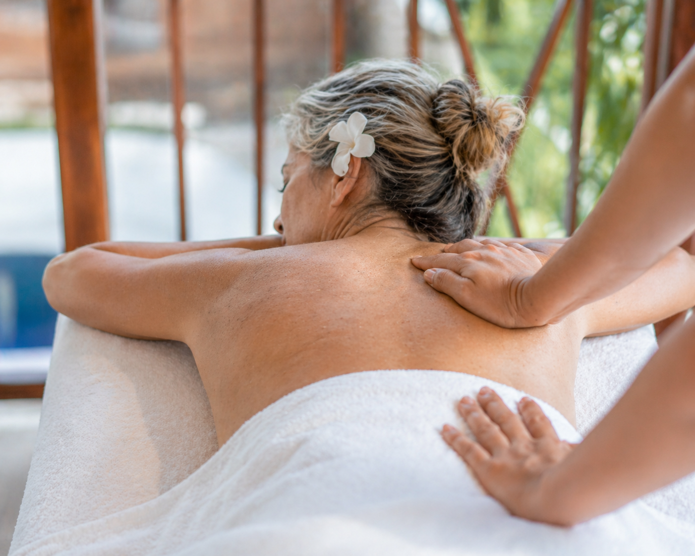 An elderly woman with gray hair and a white flower in her hair receives a back massage in a spa setting.