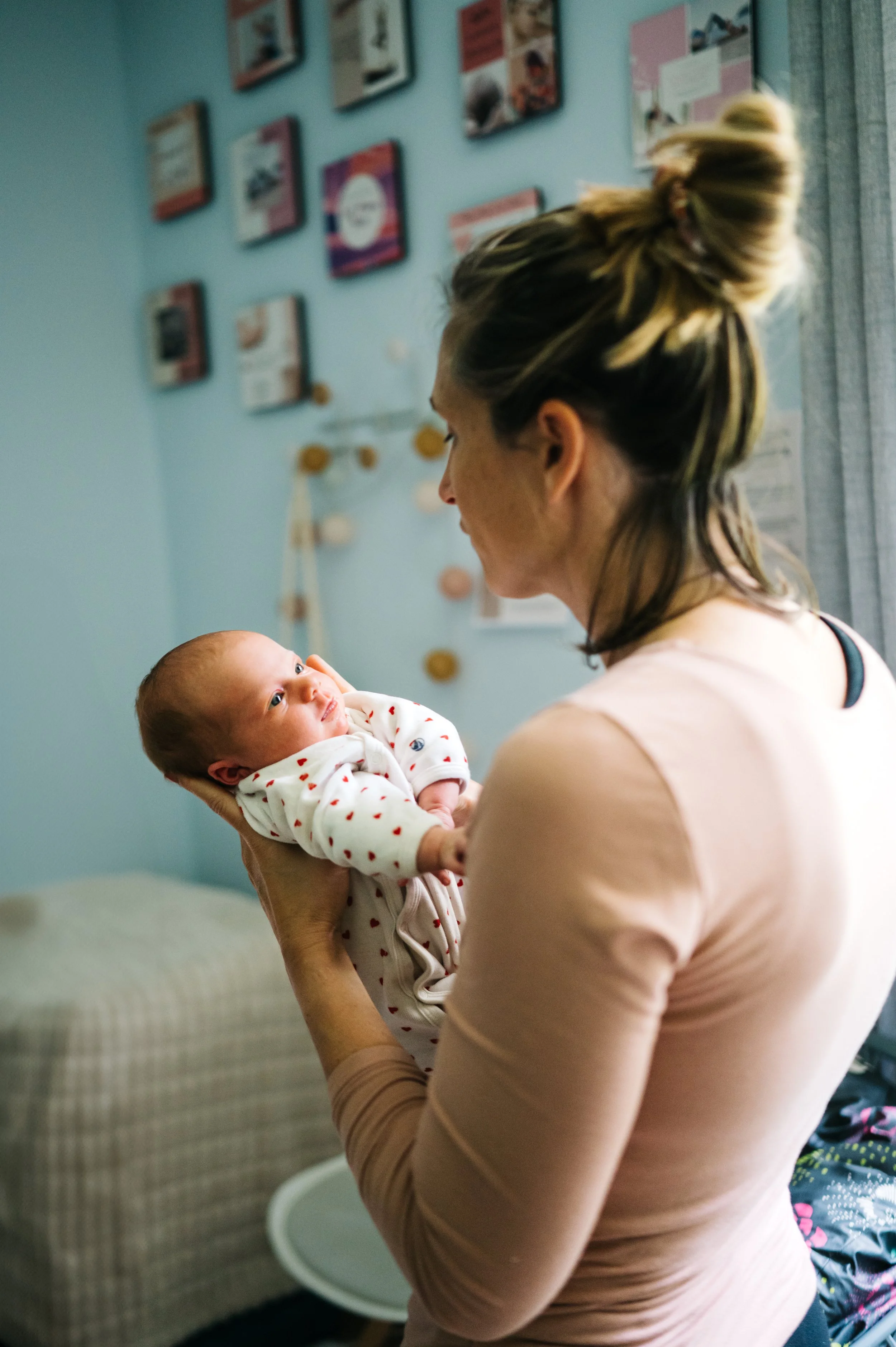 Une femme tient un bébé blanc habillé en blanc avec des petits cœurs rouges, dans une pièce avec une décoration murale colorée.