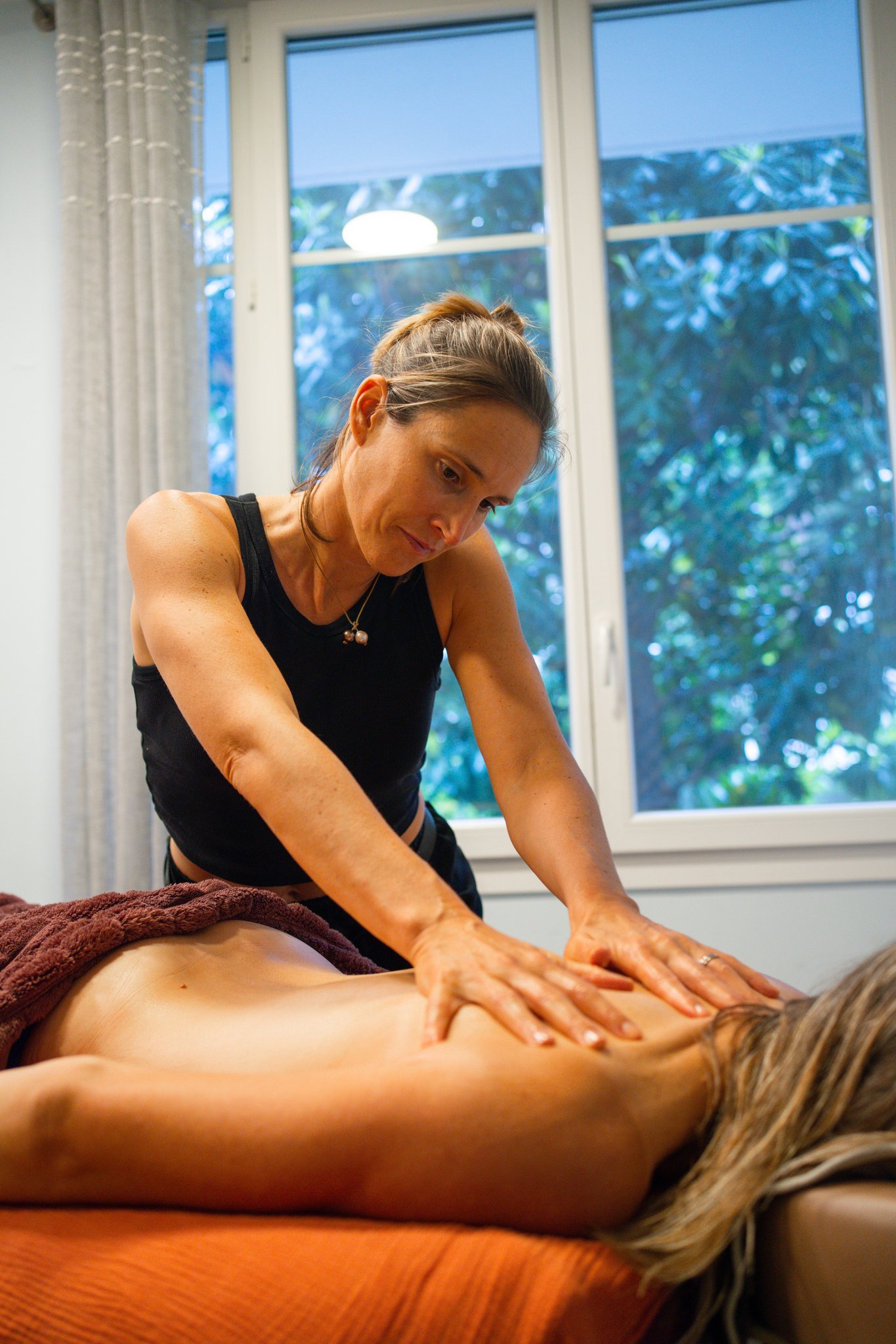 Une femme donne un massage à une personne allongée nue sur une table de massage, dans une pièce lumineuse avec vue sur des arbres à l'extérieur.