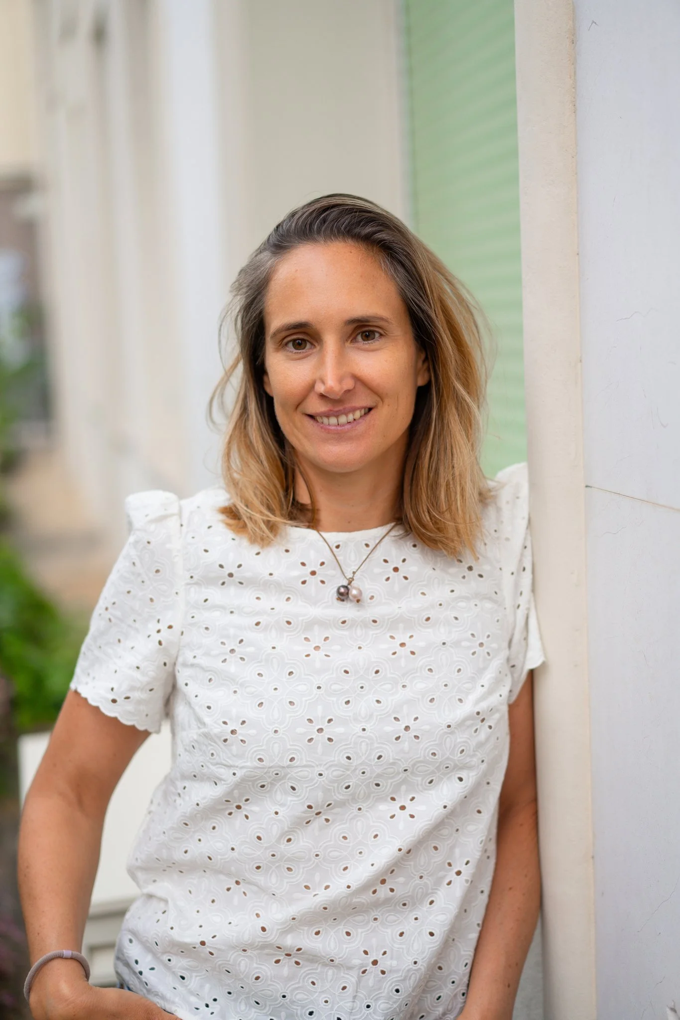 Une femme souriante à cheveux bruns côte, portant une robe blanche à motifs en broderie, avec un collier avec deux perles. Elle se tient près d’un mur blanc dans un environnement lumineux.