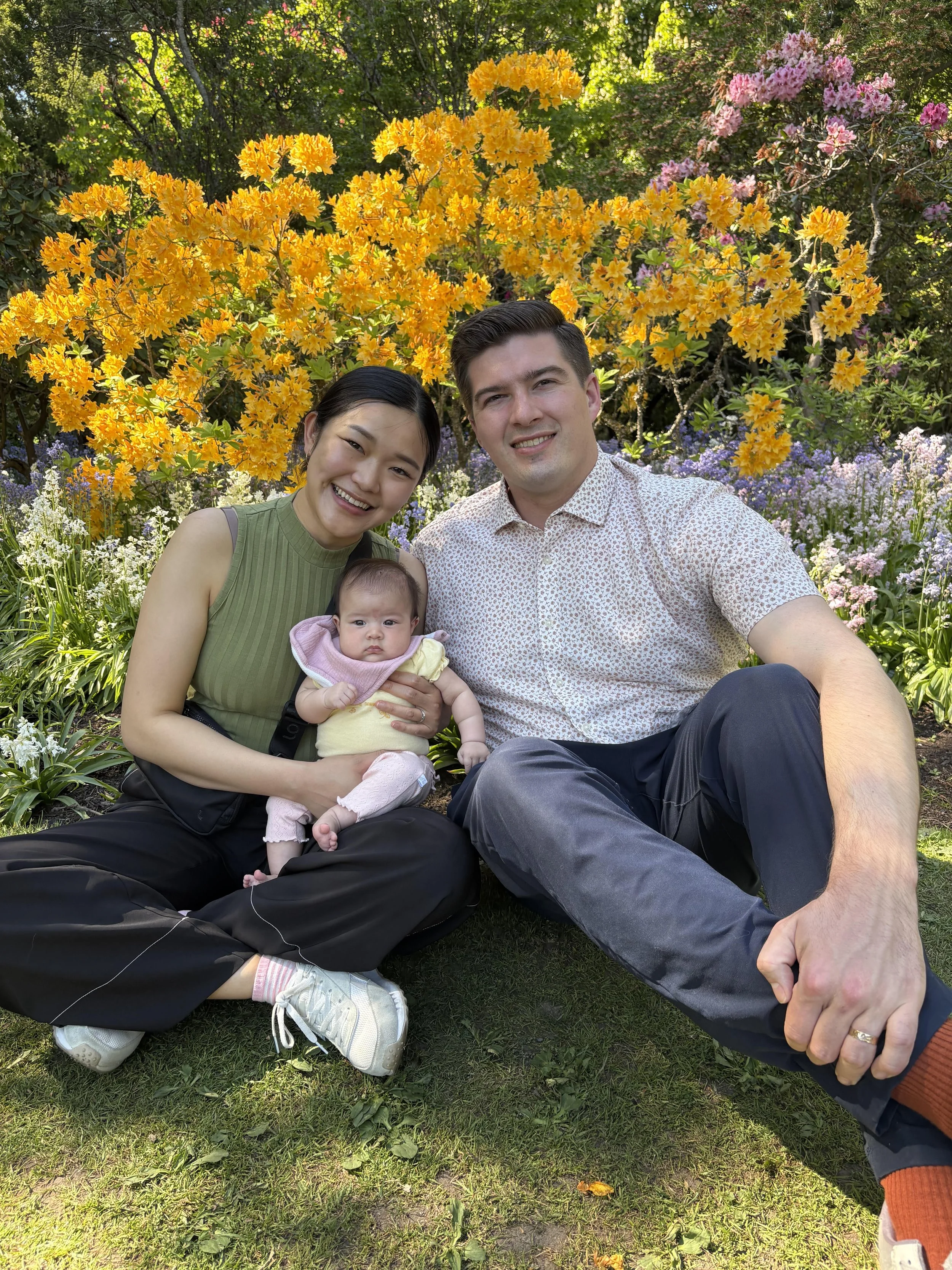 A family photo of three people, two adults and a baby, sitting outdoors in a garden with colorful flowers and greenery behind them. The woman is wearing a green top and smiling. The man is wearing a white patterned shirt and smiling. The baby is dressed in yellow and pink, looking at the camera.