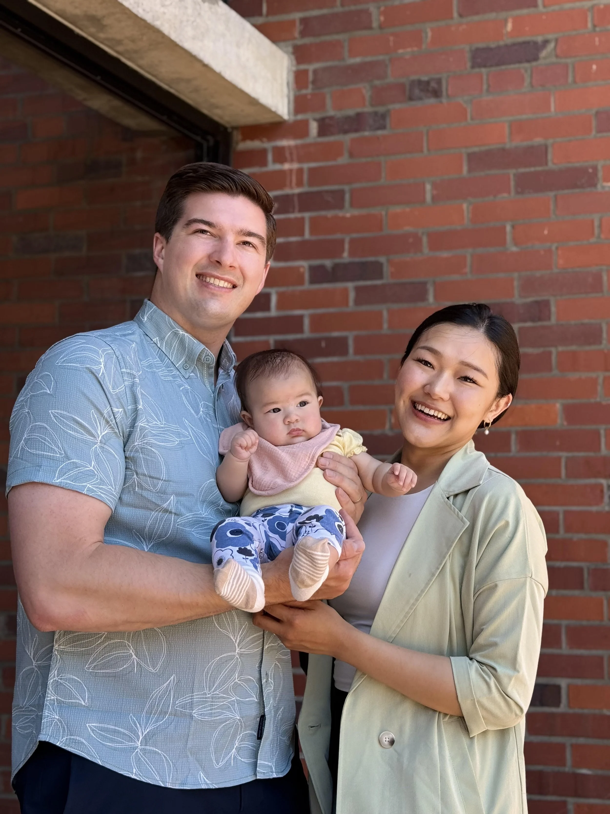 Smiling man and woman holding a baby in front of a brick wall.