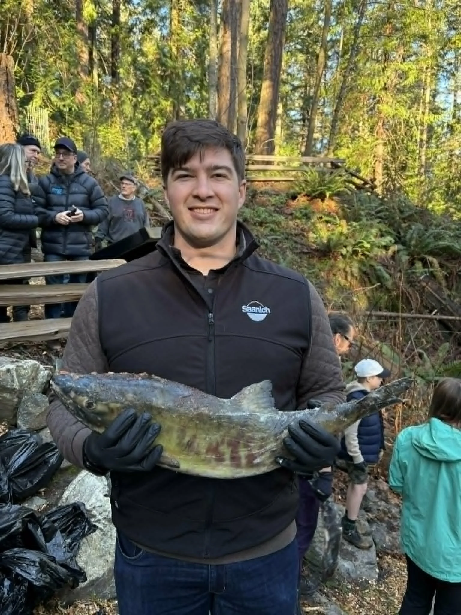 A man smiling and wearing a black jacket with a logo, holding a large fish with both hands. He is outdoors in a forested area with other people in the background.