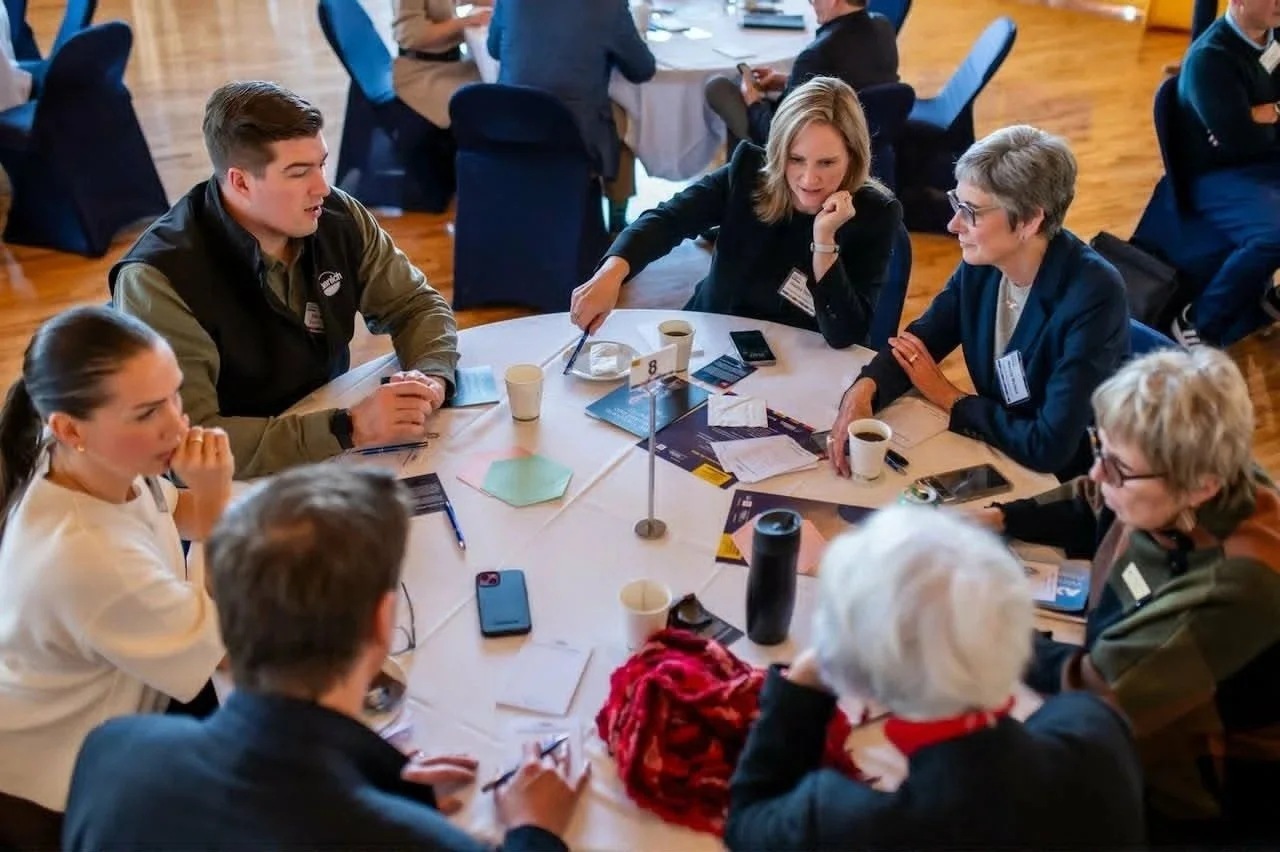 Group of people sitting around a round table, engaged in discussion, with papers, pens, cups of coffee, and electronic devices on the table, in a conference or meeting setting.
