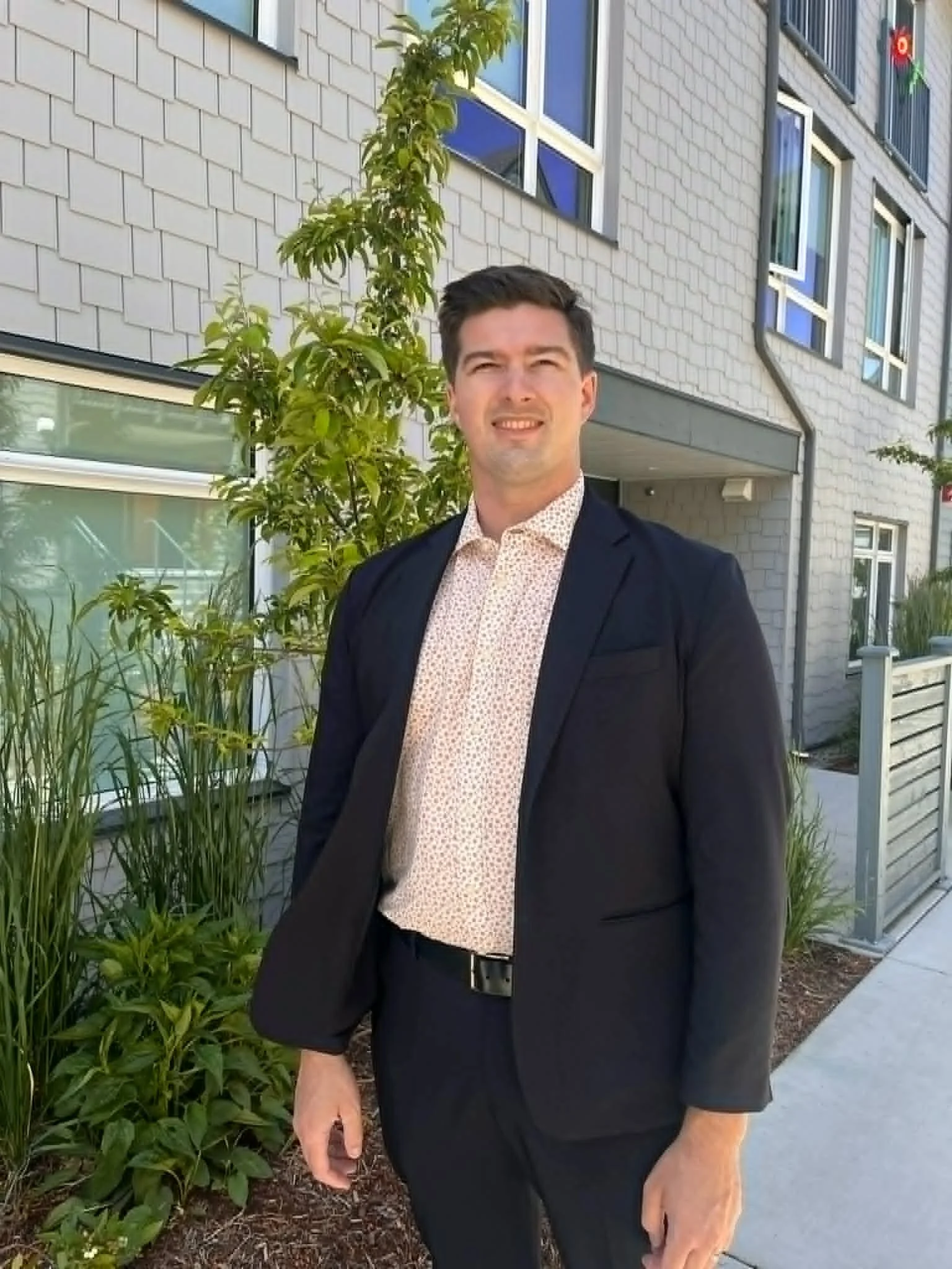A man wearing a dark suit with a white patterned shirt, standing outdoors in front of a white brick building and green plants, smiling at the camera.