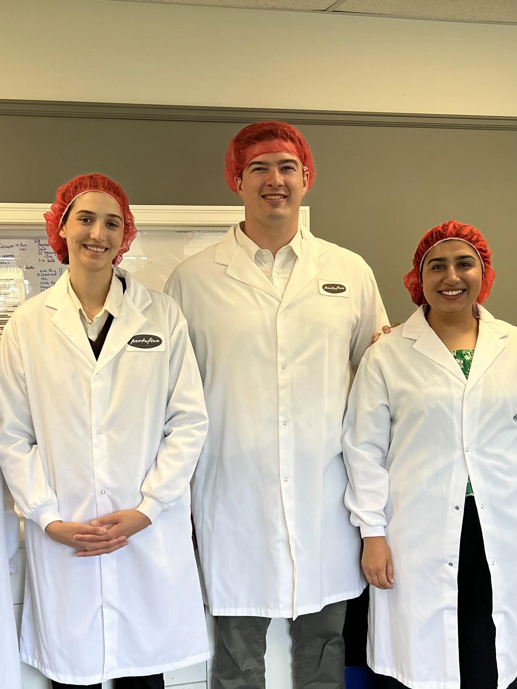 Three people wearing white coats and red hair caps, standing indoors in front of a whiteboard, smiling at the camera.