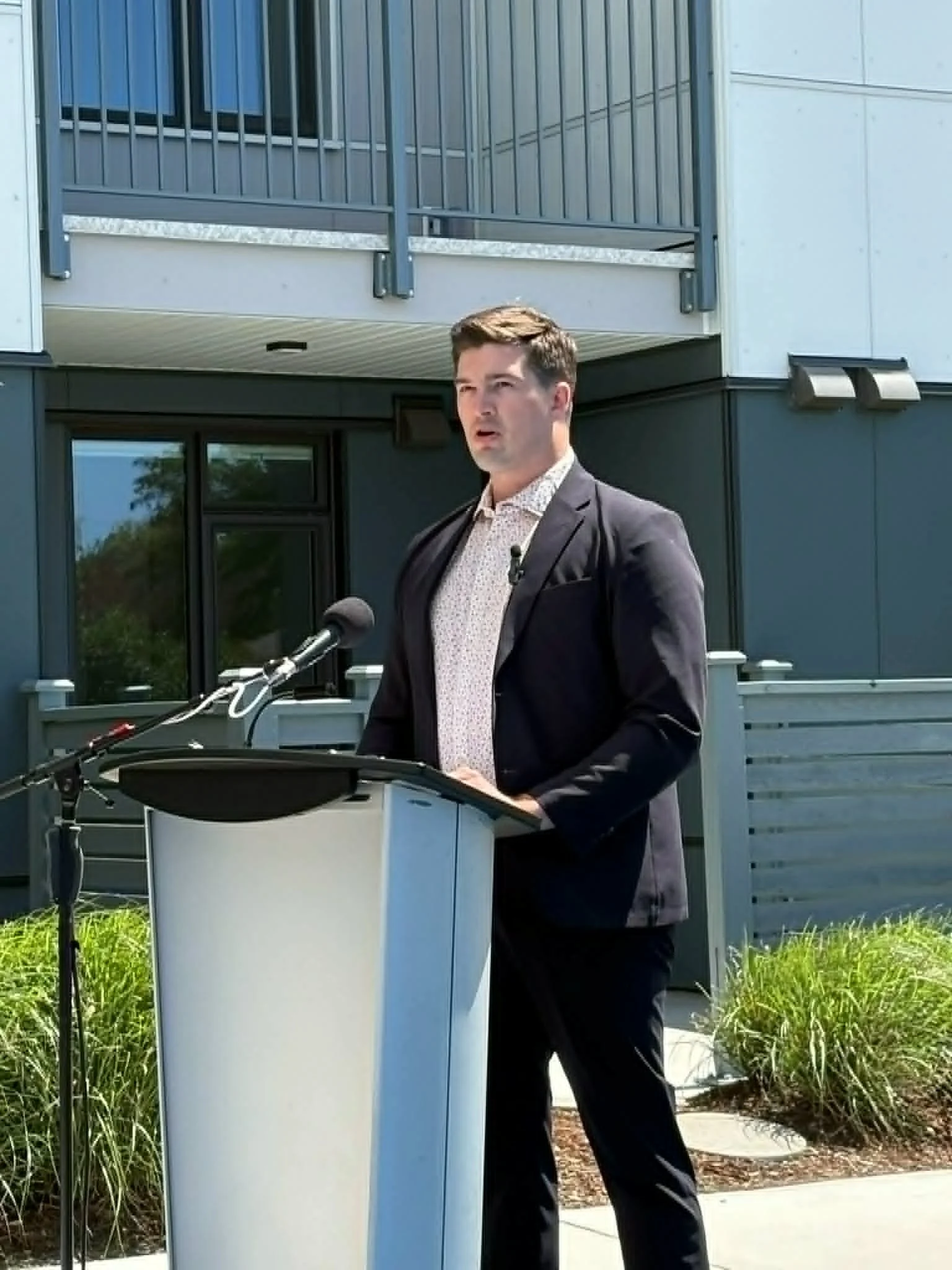 A man in a dark suit and patterned shirt giving a speech at a podium with microphones outdoors in front of a modern building with balconies.